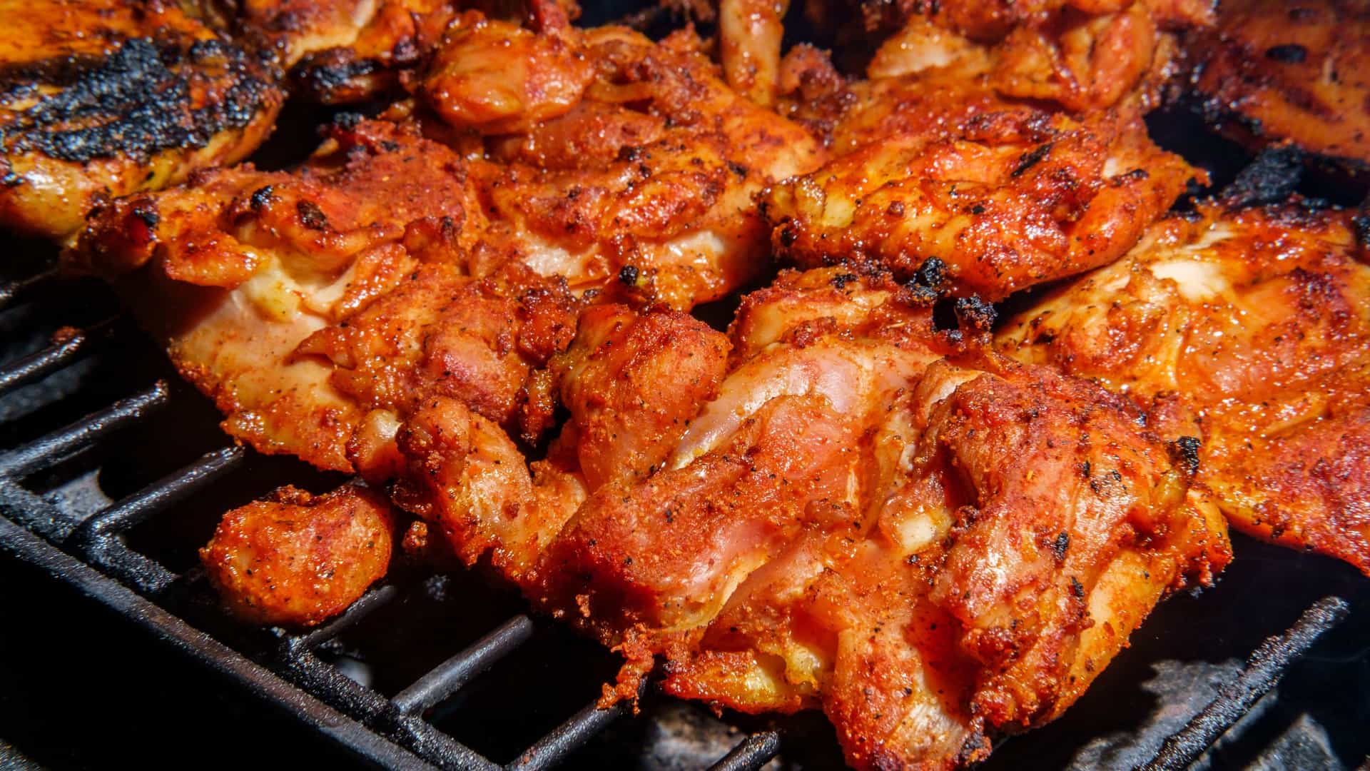 A close-up, appetizing photograph of several boneless chicken thighs sizzling directly on a hot outdoor grill grate, perfectly coated in a thick, dark reddish-brown Memphis-style dry rub seasoning. The intense heat is creating savory smoke and slight char marks on the seasoned meat.