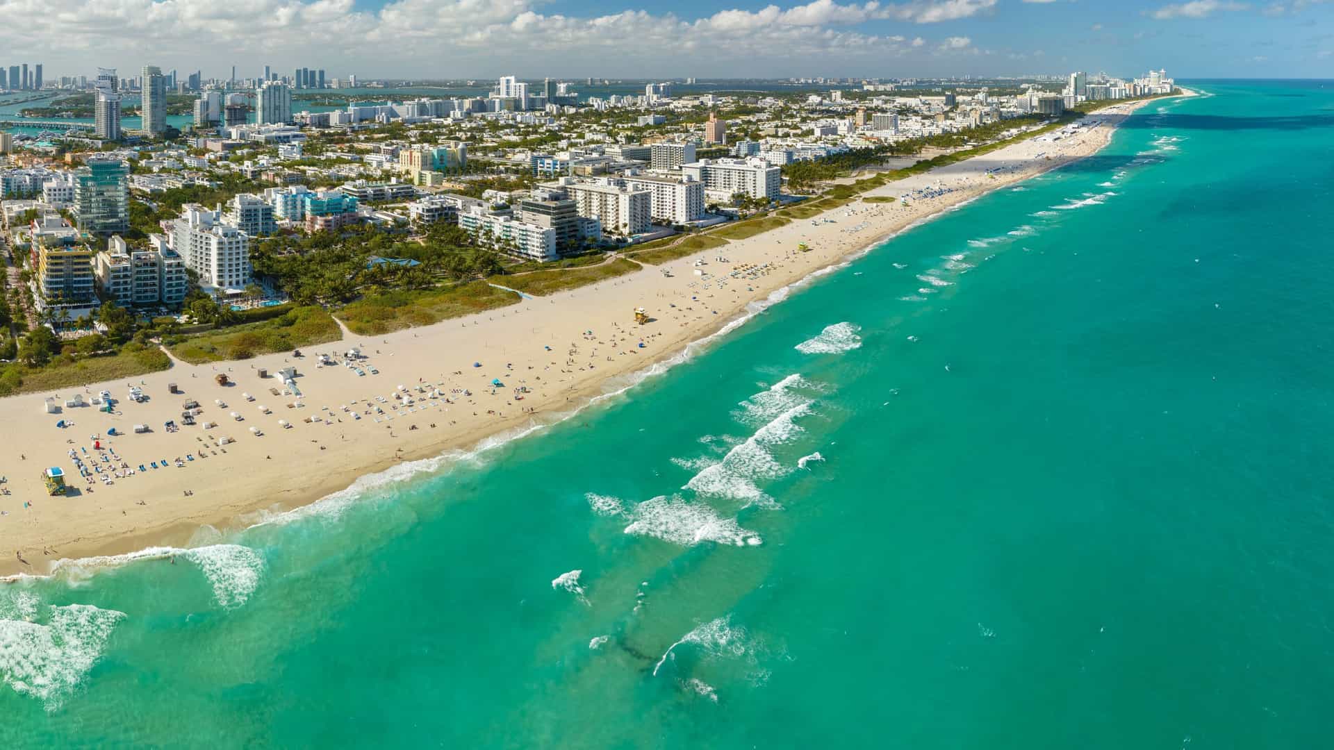 An elevated view of the famous South Beach in Miami, Florida. The image captures the wide expanse of white sand and the turquoise waters of the Atlantic Ocean. A paved beachwalk, lined with palm trees, runs parallel to the beach. In the distance, the vibrant, pastel-colored buildings of the Art Deco Historic District are visible along Ocean Drive, with the urban landscape of Miami Beach stretching out behind them.