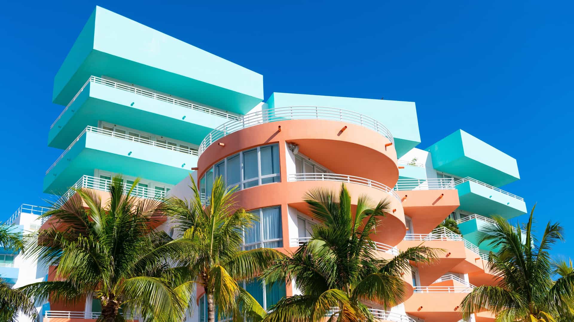 A close-up, low-angle shot of a classic Art Deco building in the Miami Beach Historic District, located on Ocean Drive in Florida. The building's facade is painted in a soft, pastel color, featuring the characteristic geometric forms, stepped-back design, and clean lines of the Art Deco style. The composition highlights the architectural details and the typical design of this famous architectural district.