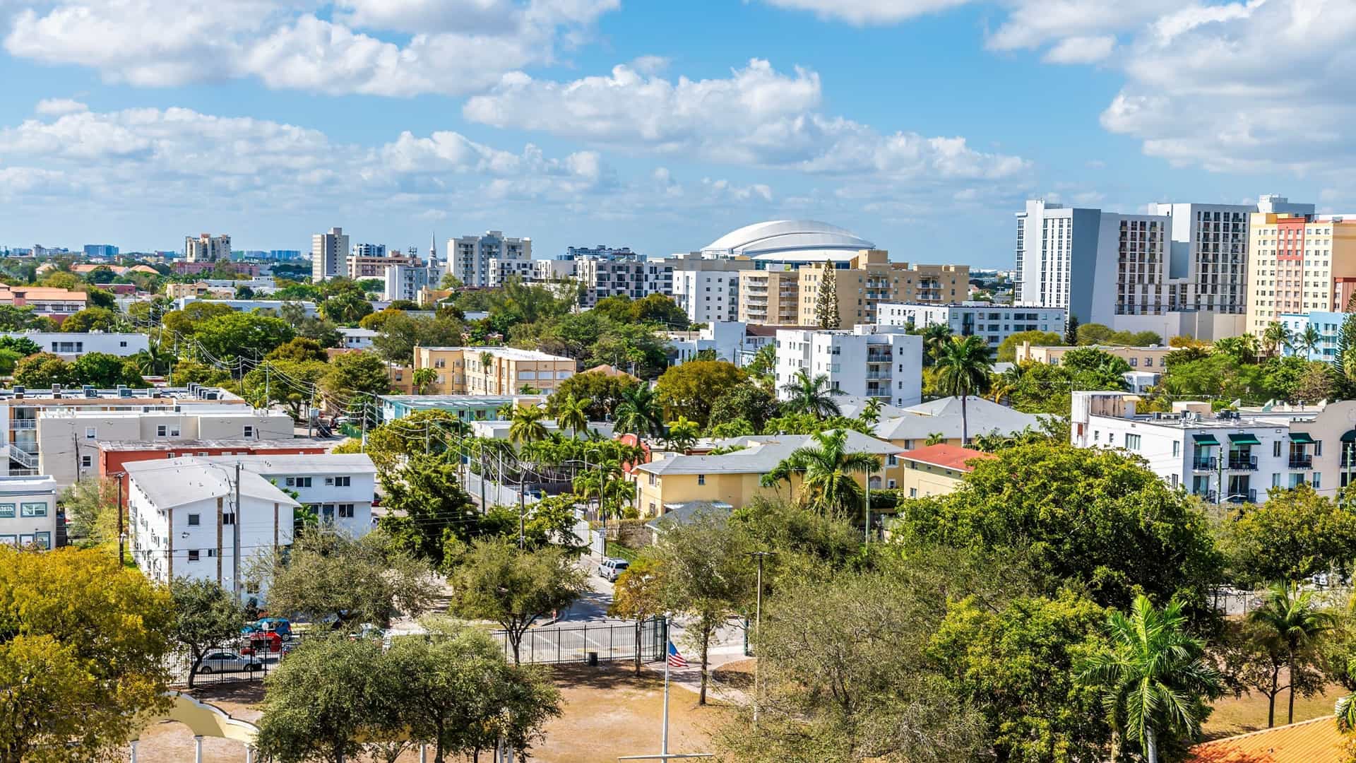 An elevated view across the Little Havana district in Miami, looking toward the modern skyline of Downtown Miami and Brickell. The foreground of the image features the lower-rise, more residential and traditional buildings of Little Havana, creating a contrast with the distant, towering skyscrapers.