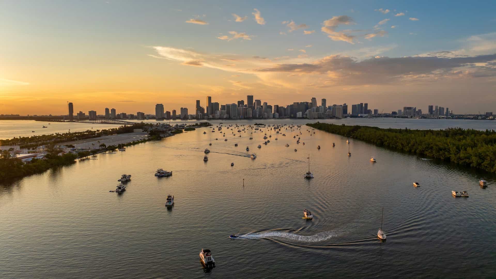A scenic view of Biscayne Bay in Miami, Florida, with a harbor filled with boats and yachts in the foreground and the modern skyline of downtown Miami and Brickell with its skyscrapers in the background under a cloudy sky.