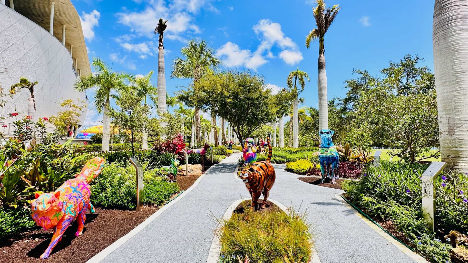 A bright, modern architectural shot of the Jorge M. Pérez Art Museum of Miami-Dade County (PAMM), featuring its distinctive hanging vertical gardens. The view captures the front facade of the building with its light-colored, tiered structure and the suspended, tropical vegetation, set against a clear sky.