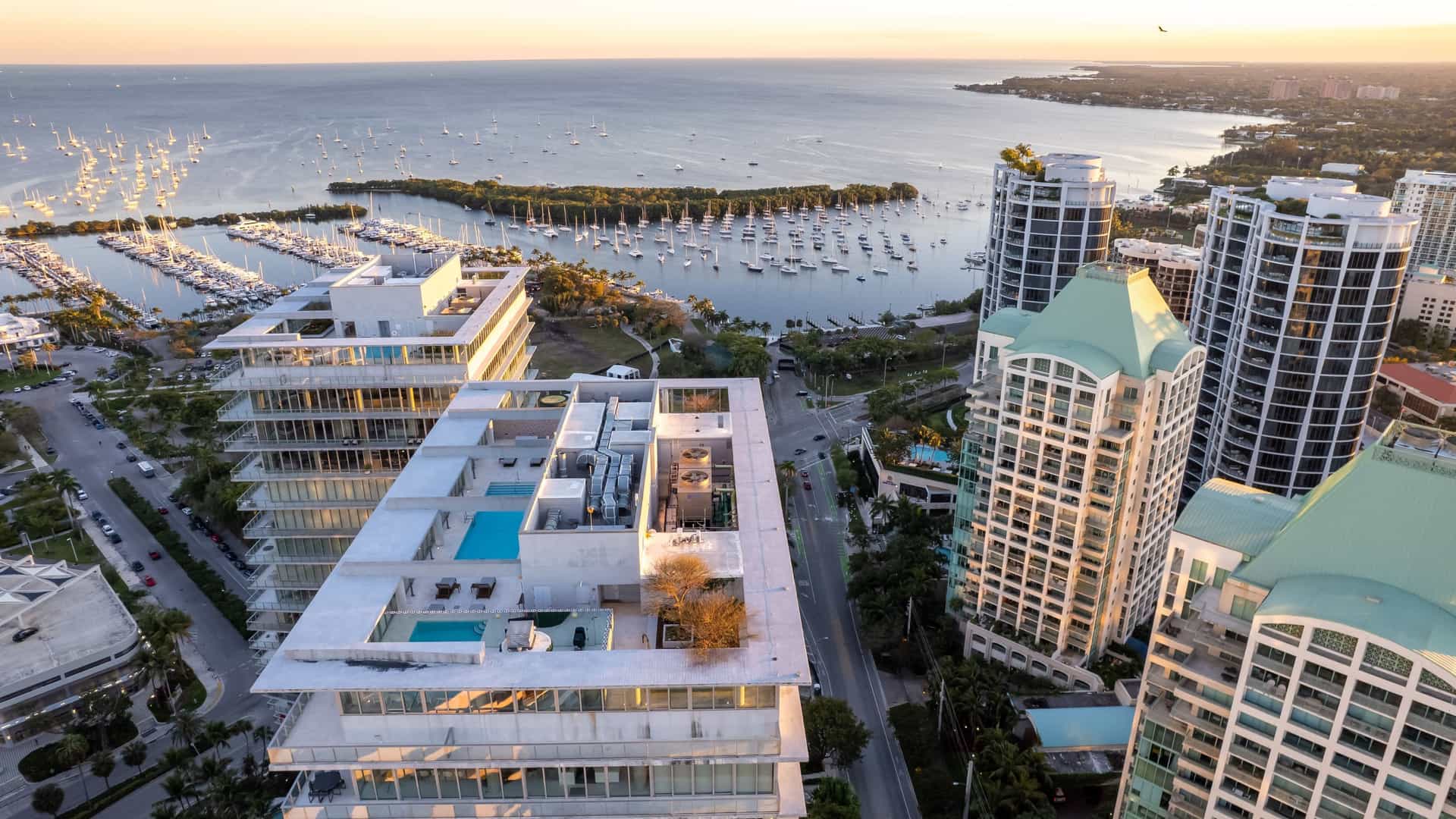 An aerial photograph of the Coconut Grove marina in Miami, Florida, captured during sunset. The image highlights the Dinner Key Marina with numerous boats and sailboats anchored in the water. The vibrant colors of the sunset, featuring warm orange and yellow tones, are reflected on the water and silhouette the palm trees along the coastline. In the background, the Miami skyline can be seen as part of the urban landscape.