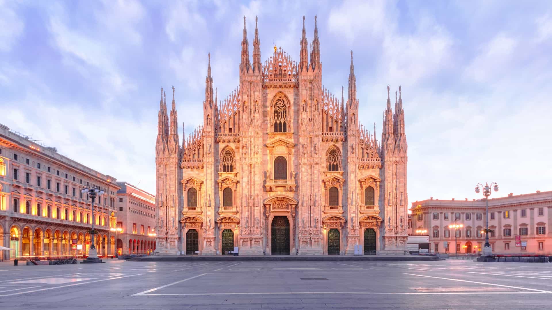 A view of the Piazza del Duomo in Milan, Italy, with the ornate, Gothic style Milan Cathedral dominating the square. The building is made of white marble and features many spires. In the foreground, people are walking and pigeons are gathered. The image captures the cathedral's intricate facade on a bright day.