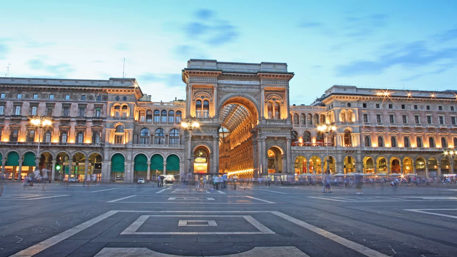 The interior of the Galleria Vittorio Emanuele II in Milan, Italy, a grand 19th century shopping arcade. The image shows the glass vaulted roof supported by an intricate cast iron framework, allowing natural light to fill the space. The elegant marble flooring and the facades of the luxury boutiques line the arcades, creating a sense of opulence.
