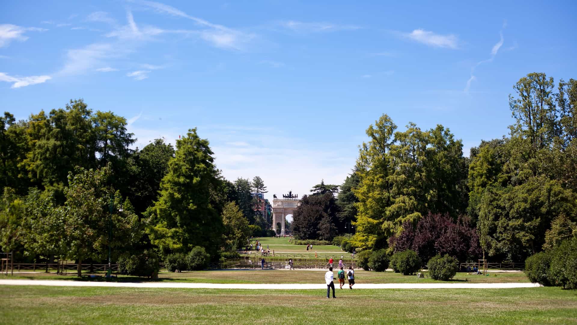 A serene view of Parco Sempione in Milan, Italy, showing a lush, green lawn bordered by a variety of trees and foliage. People are scattered across the grass, relaxing, walking, or picnicking. The image conveys a peaceful atmosphere, highlighting the park as a tranquil urban oasis.