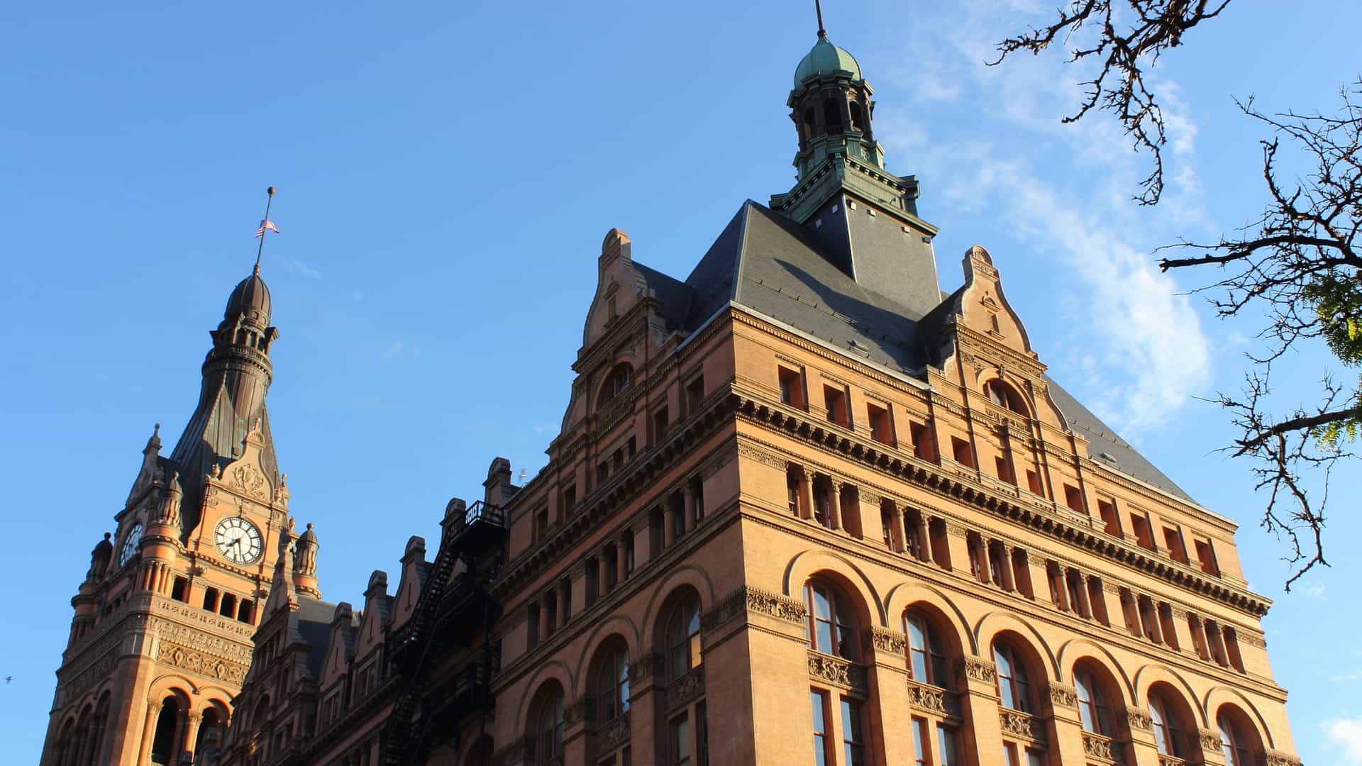 A full exterior view of the Milwaukee City Hall, a striking example of Flemish Renaissance Revival architecture. The building features light-colored brick and terra cotta, with ornate, stepped gables and a massive clock tower capped by a copper-clad spire. It is photographed from a street-level perspective, highlighting its historical grandeur against the modern cityscape.