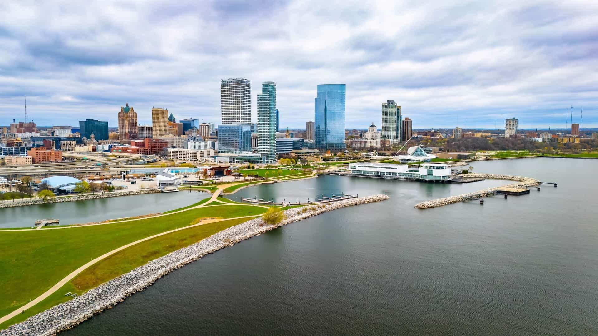 A sweeping aerial view of the Milwaukee, Wisconsin, downtown skyline situated alongside Lake Michigan under a cloudy sky. In the foreground, the modern, white Discovery World museum complex sits on a breakwater, with green parkland curving towards the tall glass skyscrapers in the background.