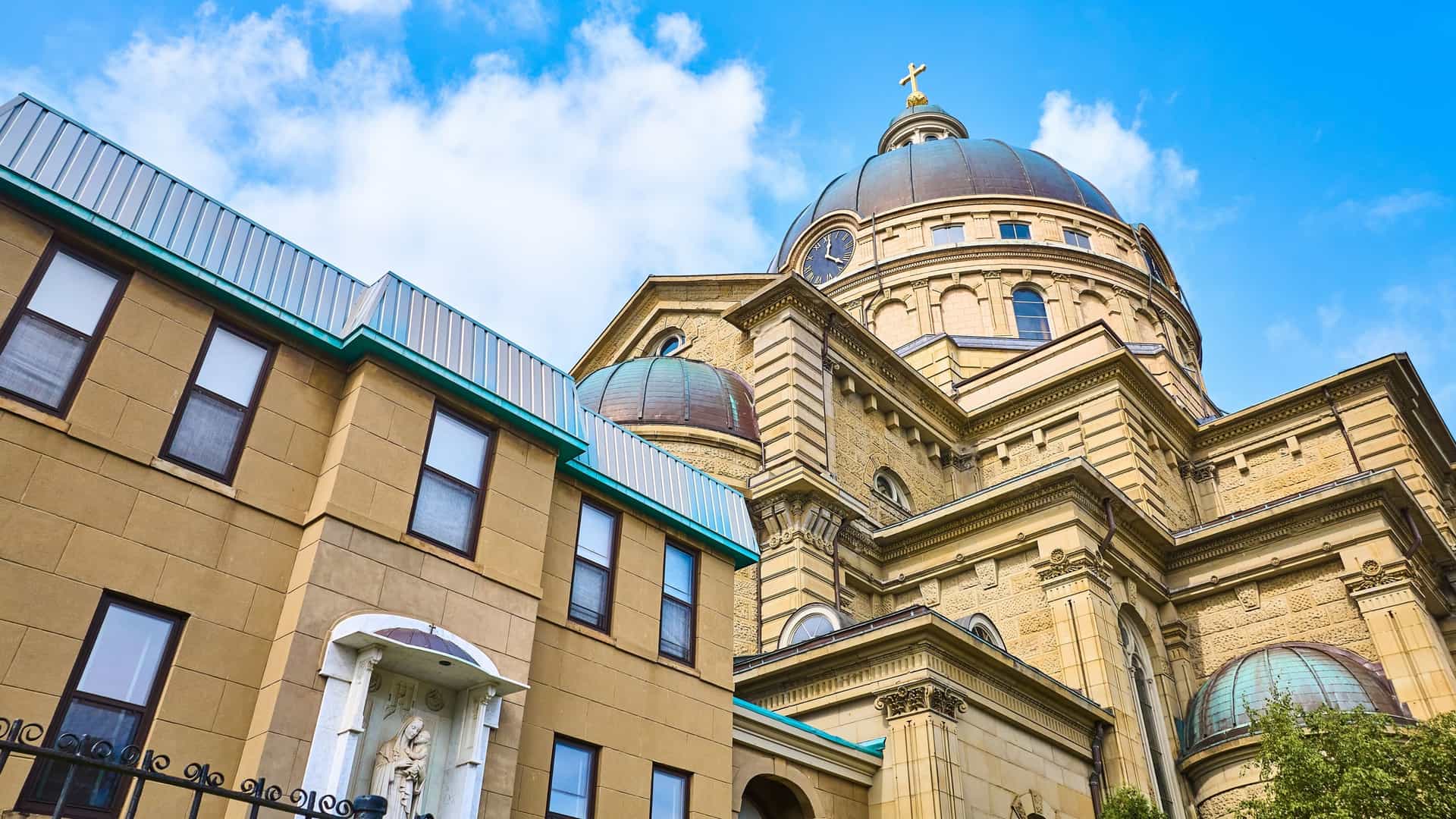 A detailed aerial close-up of the immense copper dome of the Basilica of St. Josaphat, showcasing its rich verdigris color, the ornately carved stone drum it rests on, and the bright golden cross crowning the lantern. The surrounding roof of the Basilica's transept is visible, all captured under a clear blue sky.