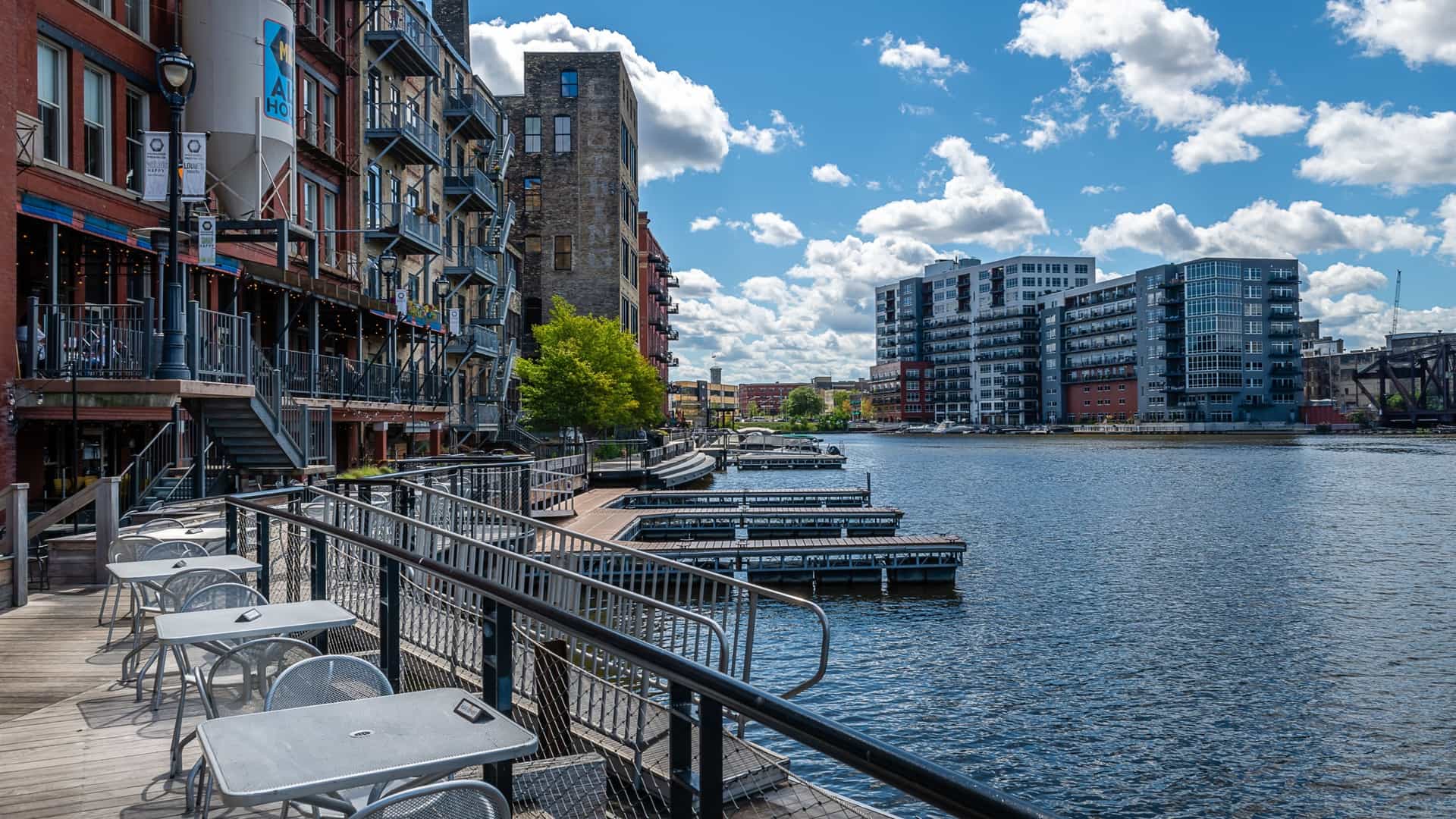A daytime photograph of the Milwaukee RiverWalk pedestrian path, showing modern and renovated historic buildings lining the river. The walkway features benches, landscaping, and railing, with the calm river water reflecting the architecture of the downtown district.