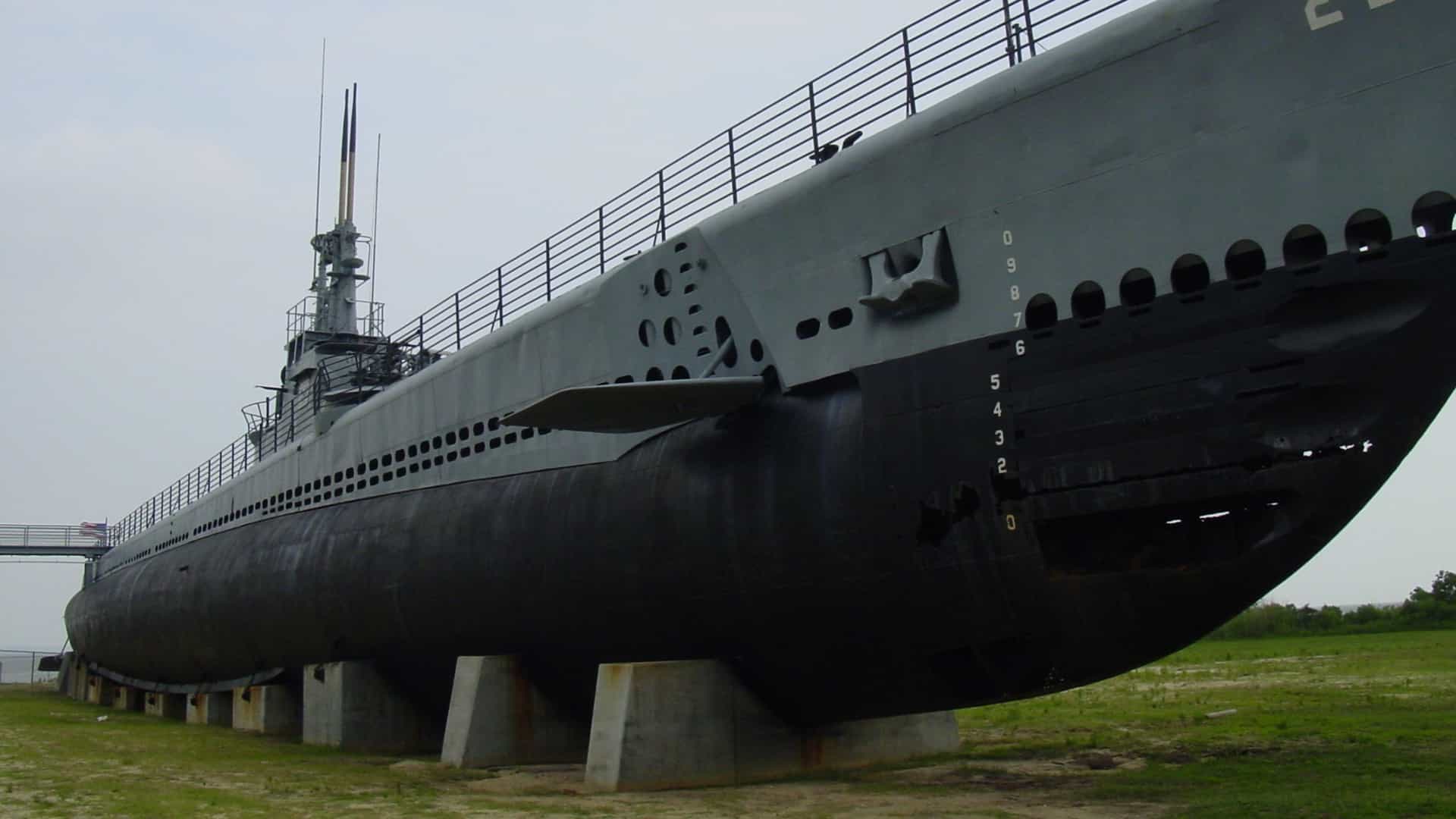 A low-angle, exterior photograph of the USS Drum (SS-228), a historic Gato-class submarine and National Historic Landmark, displayed permanently on dry land at the USS Alabama Battleship Memorial Park in Mobile, Alabama. The massive hull rests on concrete cradles, with the conning tower and the hull number "228" clearly visible under an overcast sky.