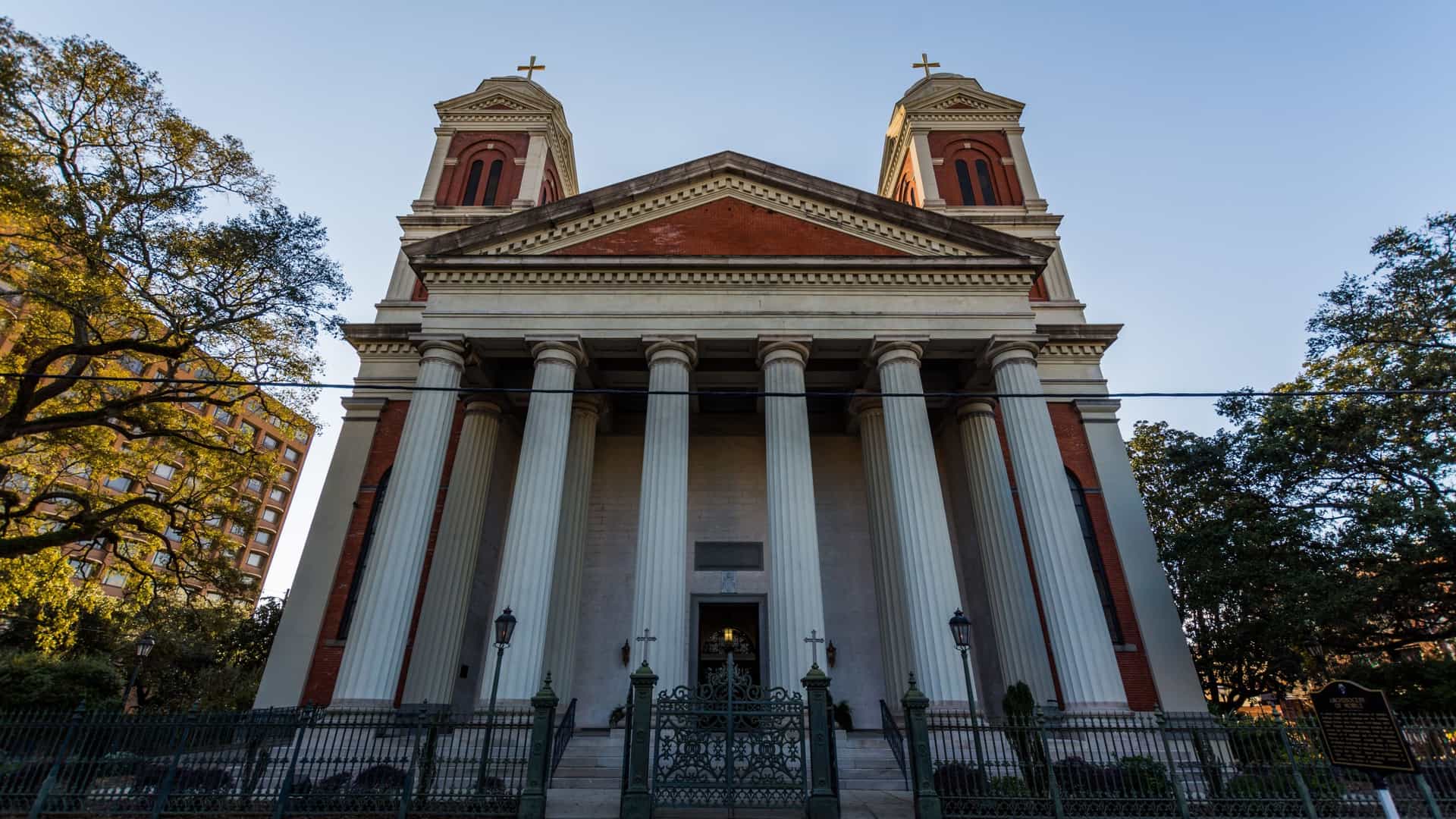 The exterior view of the historic Cathedral Basilica of the Immaculate Conception in Mobile, Alabama. The structure features a large, dominant Roman basilica-style portico with four massive columns supporting a triangular pediment. Twin, square bell towers topped with steeples flank the main entrance, and the entire front is framed by a decorative black wrought-iron fence.