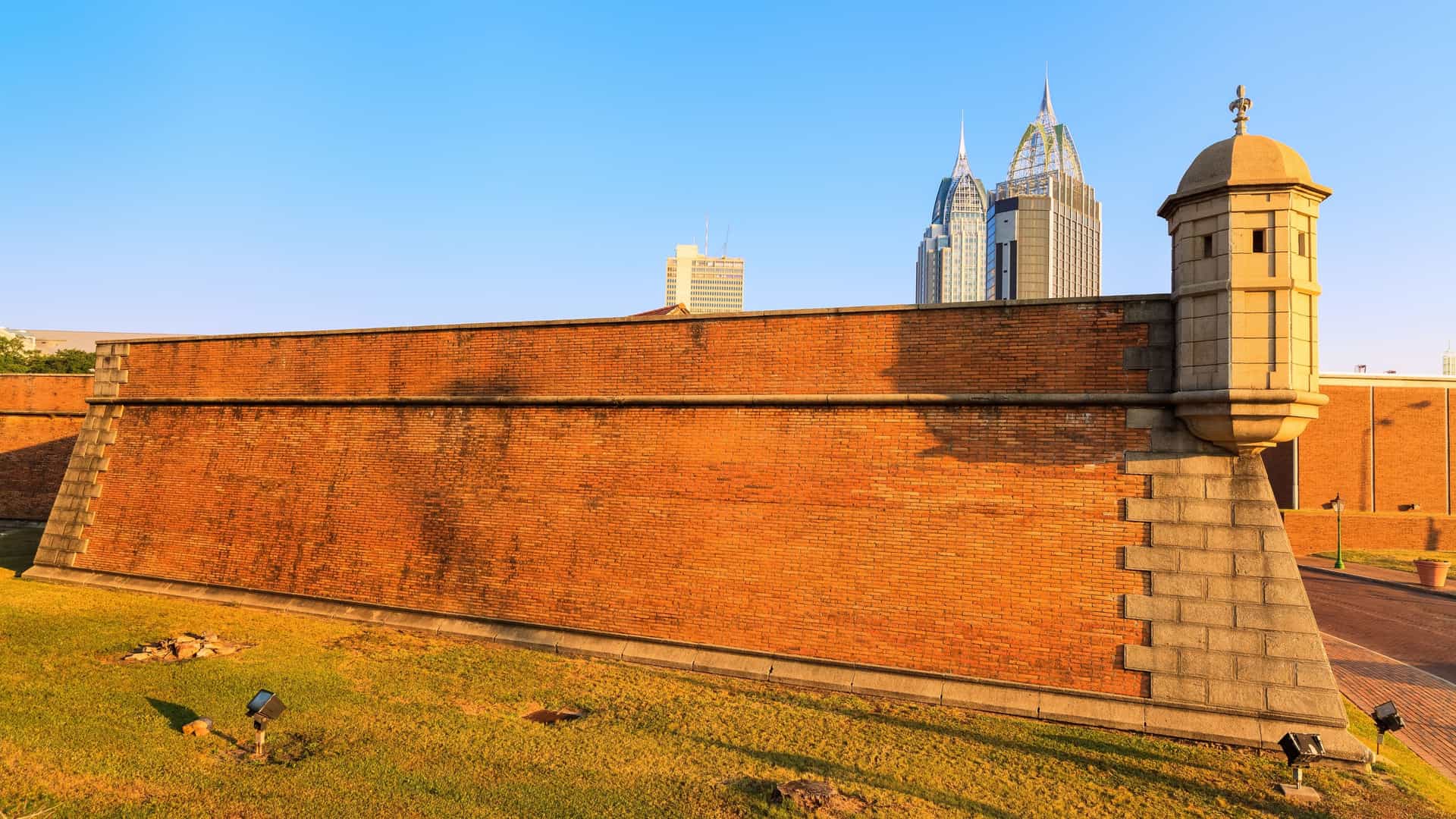 A low-angle exterior view of the Colonial Fort Condé reconstruction in Mobile, Alabama. The main gate and high brick walls of the fort are visible, topped by wooden ramparts and a sentry box. The modern downtown Mobile skyline, including tall glass and concrete buildings, is visible rising sharply behind the historic-style brick structure.