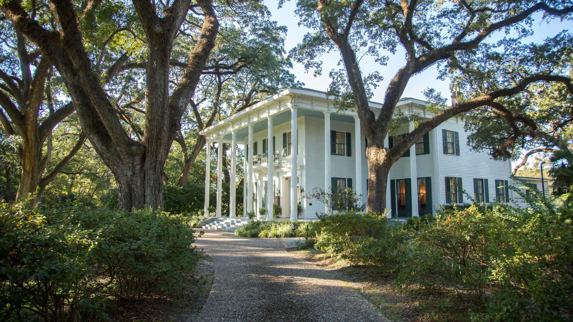 The Bragg-Mitchell Mansion, a historic white Greek Revival home in Mobile, Alabama, viewed from the end of a gravel path. The two-story structure features a grand facade with tall white columns supporting a double veranda. The mansion is heavily framed by the dark, gnarled trunks and spreading canopies of large live oak trees, with lush green bushes lining the path.