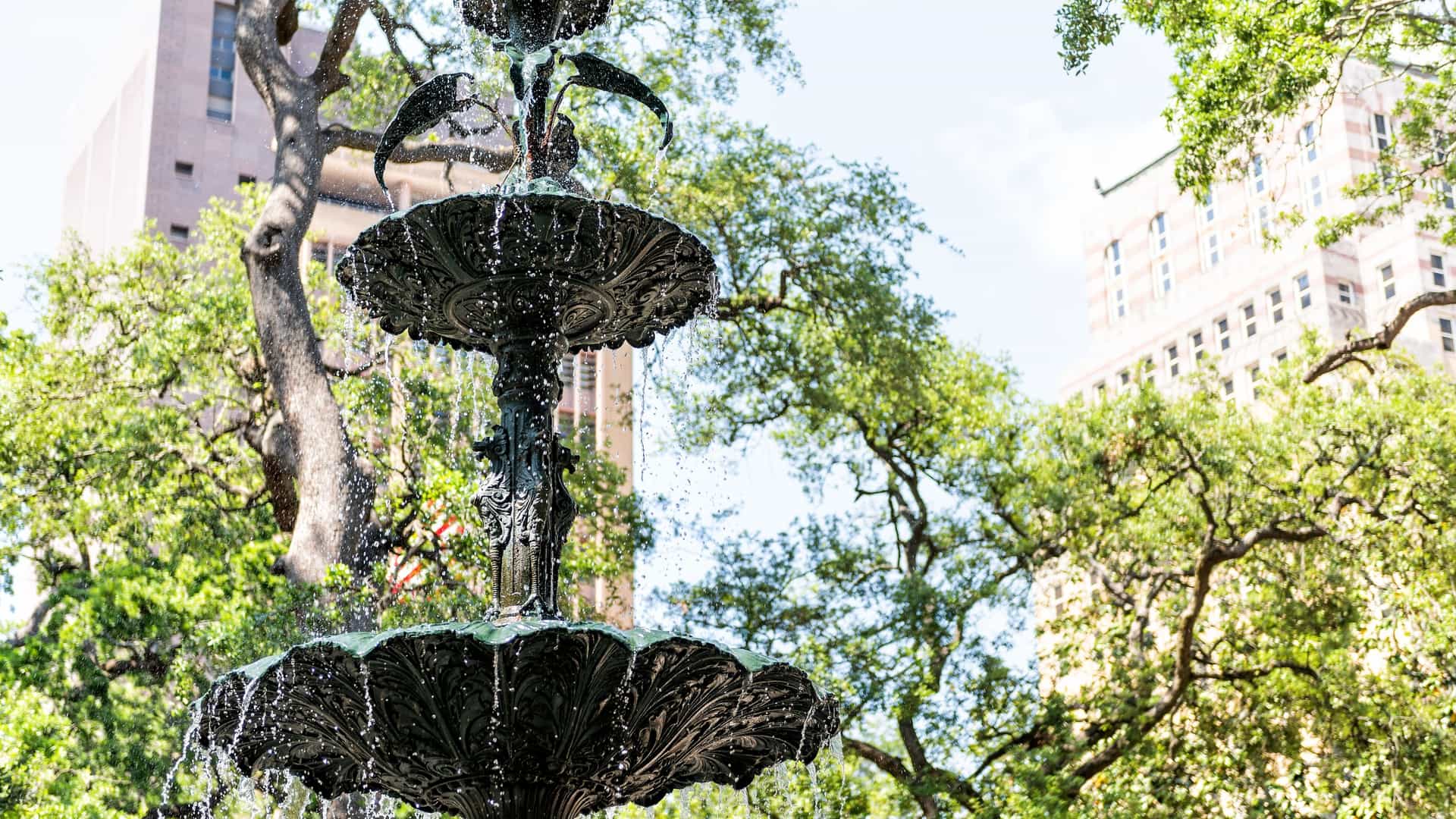 A sunny wide-angle view of Bienville Square, a historic public park in downtown Mobile, Alabama. The scene is dominated by the ornate, multi-tiered cast-iron Ketchum Fountain in the center of a paved plaza. The square is surrounded by mature live oak trees and historic downtown buildings, with people and vehicles visible on the surrounding street.