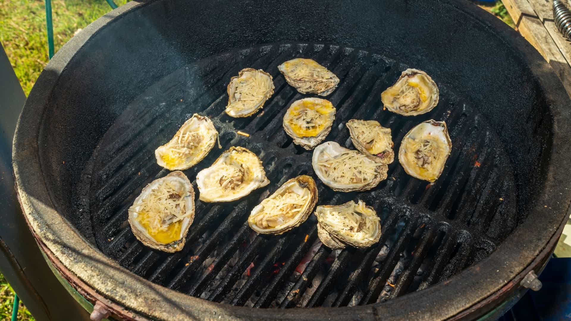 A close-up, high-angle view of a dozen or more fresh oysters grilling in their shells on a metal grate. The oysters are topped with melted butter, cheese, and herbs, and are bubbling over the hot coals, creating a smoky, savory effect.
