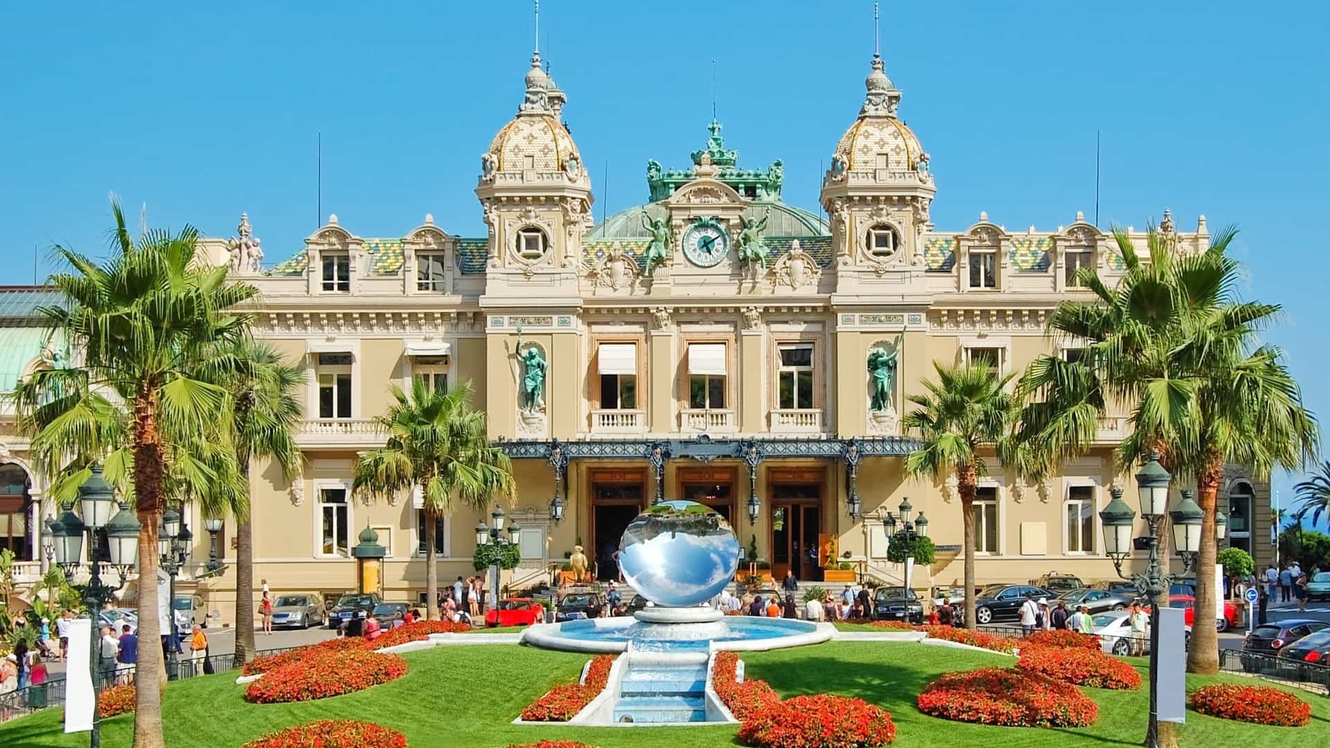 A front view of the Grand Casino Monte Carlo in Monaco, a lavishly decorated building in the Belle Époque architectural style. The ornate facade features a large clock flanked by sculptures, with intricate detailing and decorative elements. The building stands on a terrace with manicured gardens, and in front, a circular plaza with trees and streetlights is visible. The image captures the casino's opulent and historic appearance.