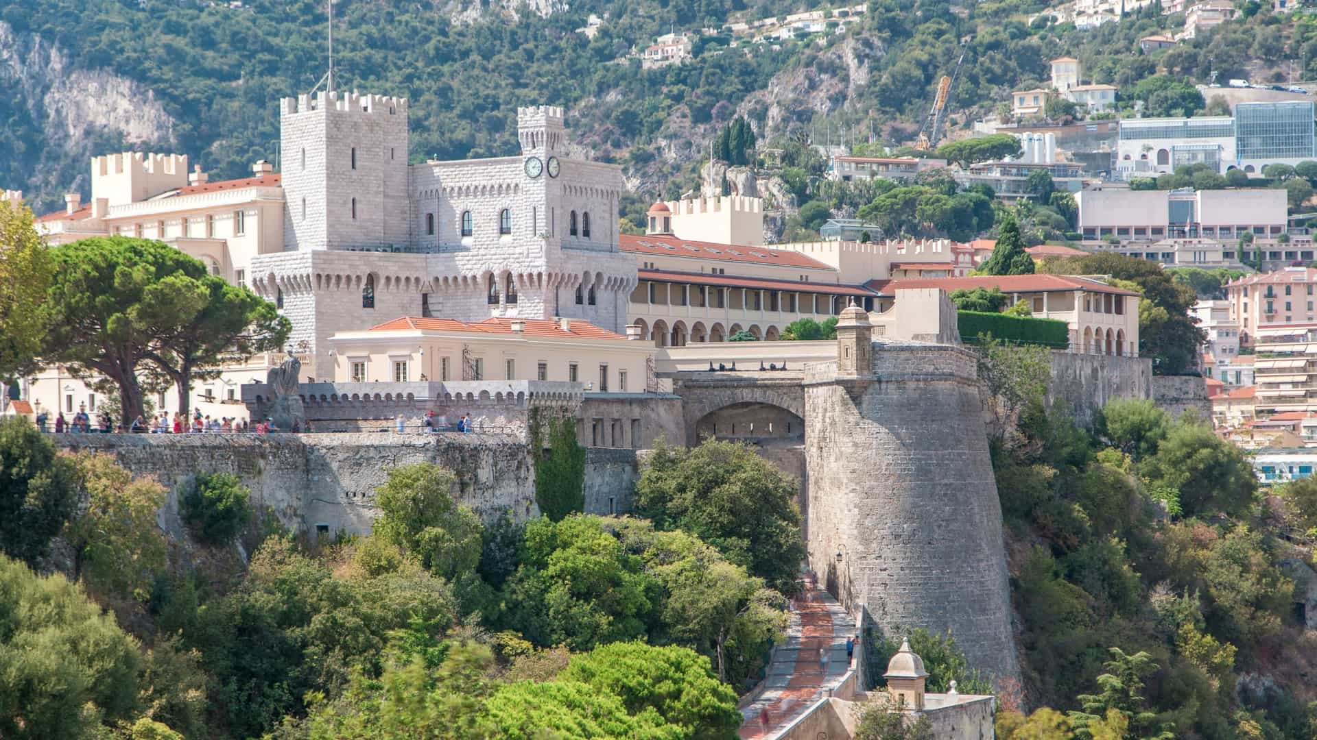 A bright, sunny daytime shot of the Prince's Palace of Monaco, the official residence of the Prince of Monaco. The image is taken from an observation deck, providing a view of the palace's facade and the surrounding historic buildings of Monaco Ville. The clear blue sky and warm lighting indicate it's a sunny summer day.