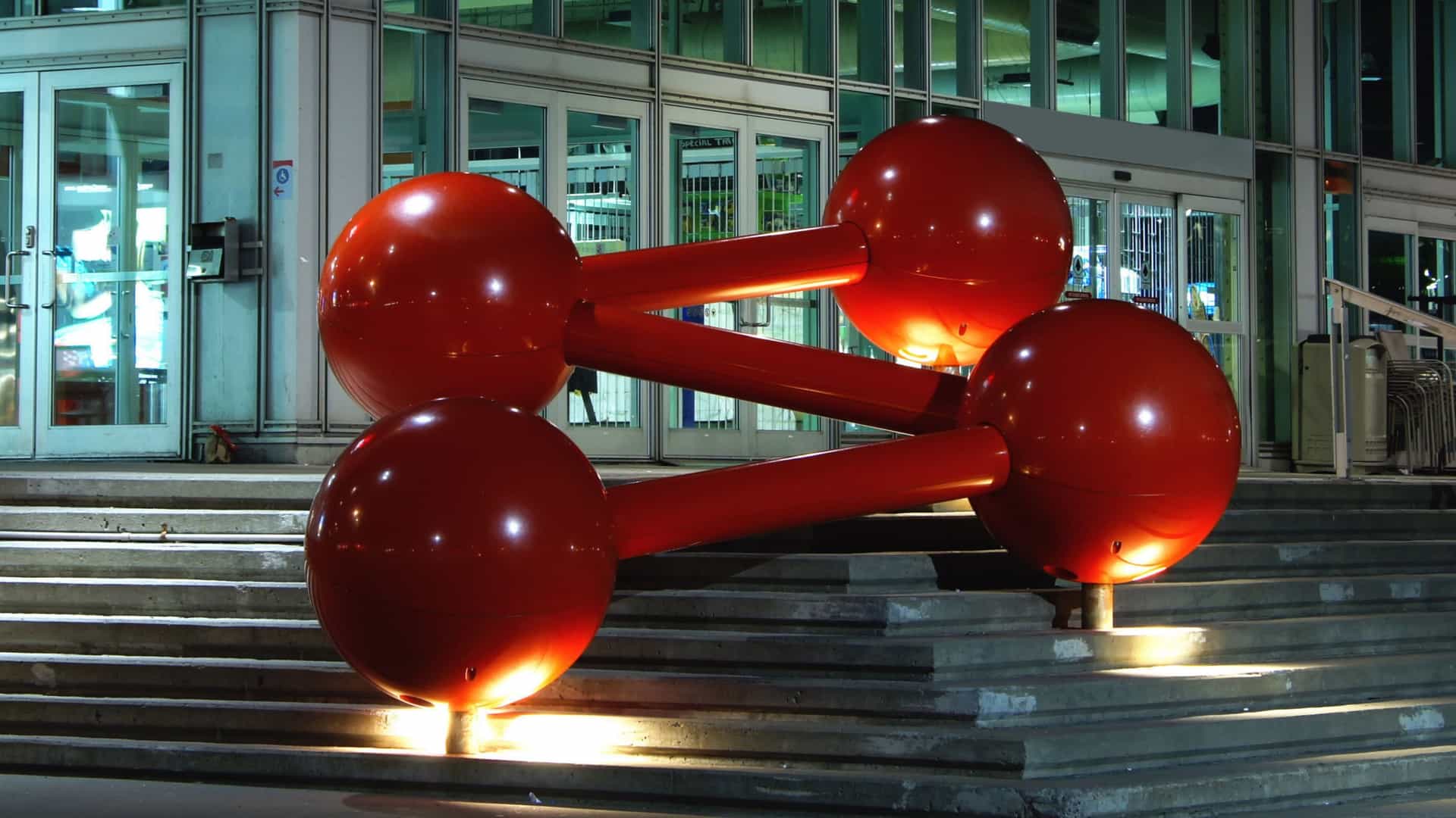 A night-time view of the modern, glass-and-steel facade of the Montréal Science Centre (Centre des sciences de Montréal) at the Old Port. The building features large, multi-story glass panels. The foreground is dominated by a prominent, circular structure—a stylized, wireframe globe or atomic symbol—which is brightly illuminated in blue light, serving as a landmark or symbol for the science center.