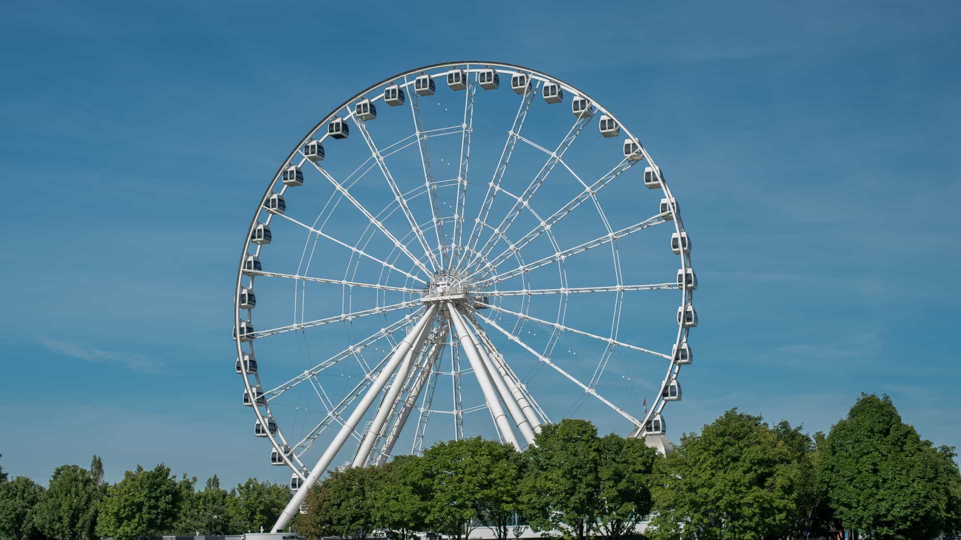 An aerial or elevated view of La Grande Roue de Montréal (The Montreal Observation Wheel) and the Old Port of Montreal, situated on the Bonsecours Basin Island along the St. Lawrence River. The Ferris wheel, the tallest in Canada at 60 meters, dominates the frame with its white structure and numerous enclosed, climate-controlled cabins. In the background, the historic red-brick buildings of Old Montreal and the modern skyline of the city rise up, with the river visible in the foreground.