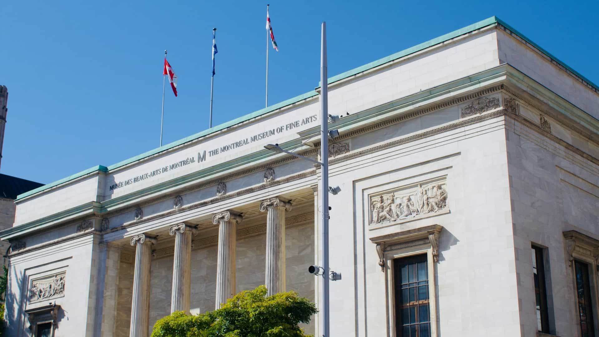 A detailed photograph of the contemporary entrance to the Montreal Museum of Fine Arts, showcasing the striking, multi-story glass canopy of the Jean-Noël Desmarais Pavilion. The modern facade features expansive transparent glass walls set against polished white marble cladding, creating a luminous and architecturally significant grand entrance to the renowned museum complex.