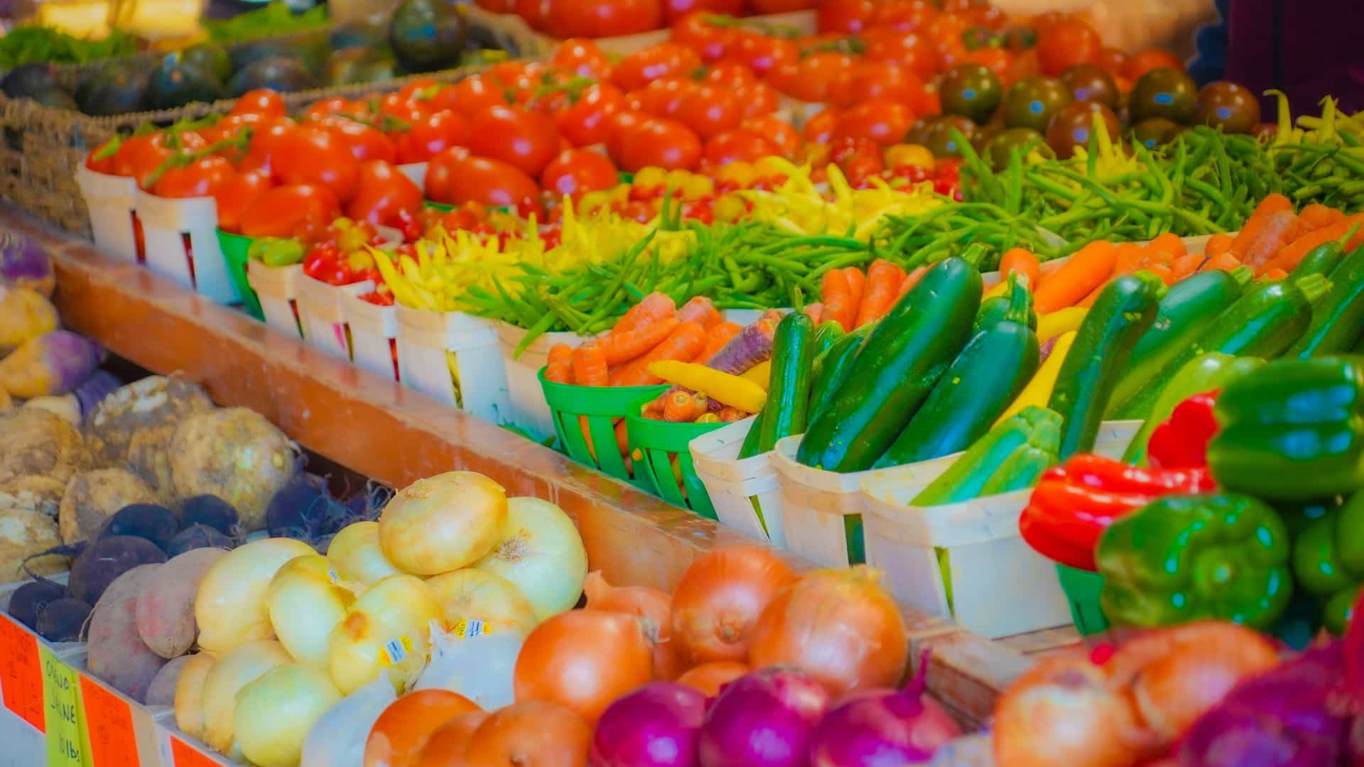 A vibrant, close-up photograph capturing the abundant fresh produce displays at Montreal's renowned Jean-Talon Market, one of North America's largest open-air markets. Rows of colorful, high-quality seasonal vegetables, including various squashes, cabbages, and root vegetables, are neatly arranged on vendor stalls under a large, covered market roof, conveying a lively harvest scene.