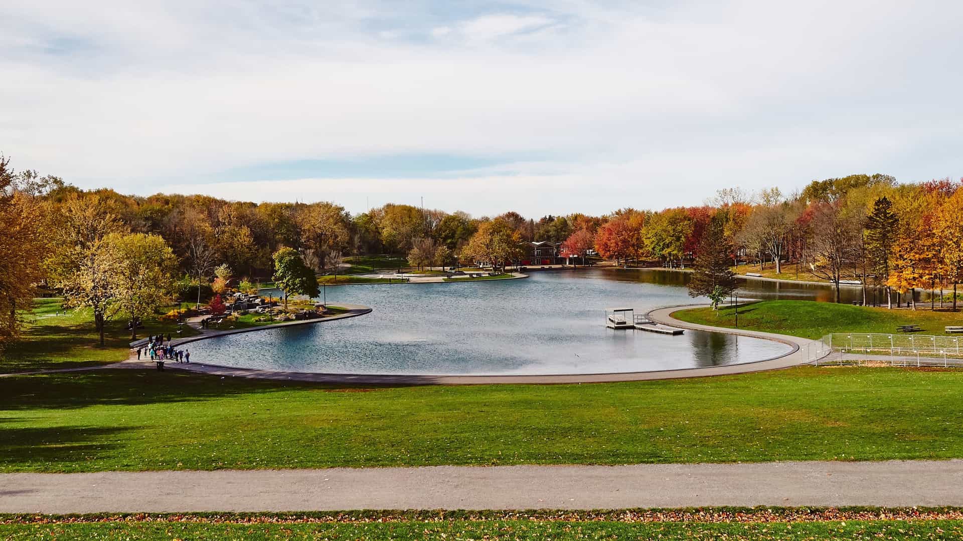 A serene view of Beaver Lake (Lac aux Castors) in Mount Royal Park, Montreal, showing the calm, reflective water surrounded by a variety of green trees and an open, grassy area, with a paved walkway in the foreground and a modern, low lying pavilion building in the distance. The sky is bright and a few people can be seen relaxing in the park.