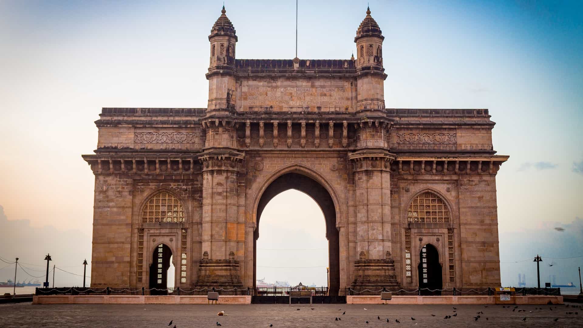 A close up view of the Gateway of India, a basalt arch monument in Mumbai, India, with its detailed Indo Saracenic architecture visible, including the intricate latticework and the large central dome. In the background, the iconic Taj Mahal Palace and Tower Hotel is visible, and the monument is surrounded by a crowd of tourists on the waterfront. The sky is bright and slightly hazy.