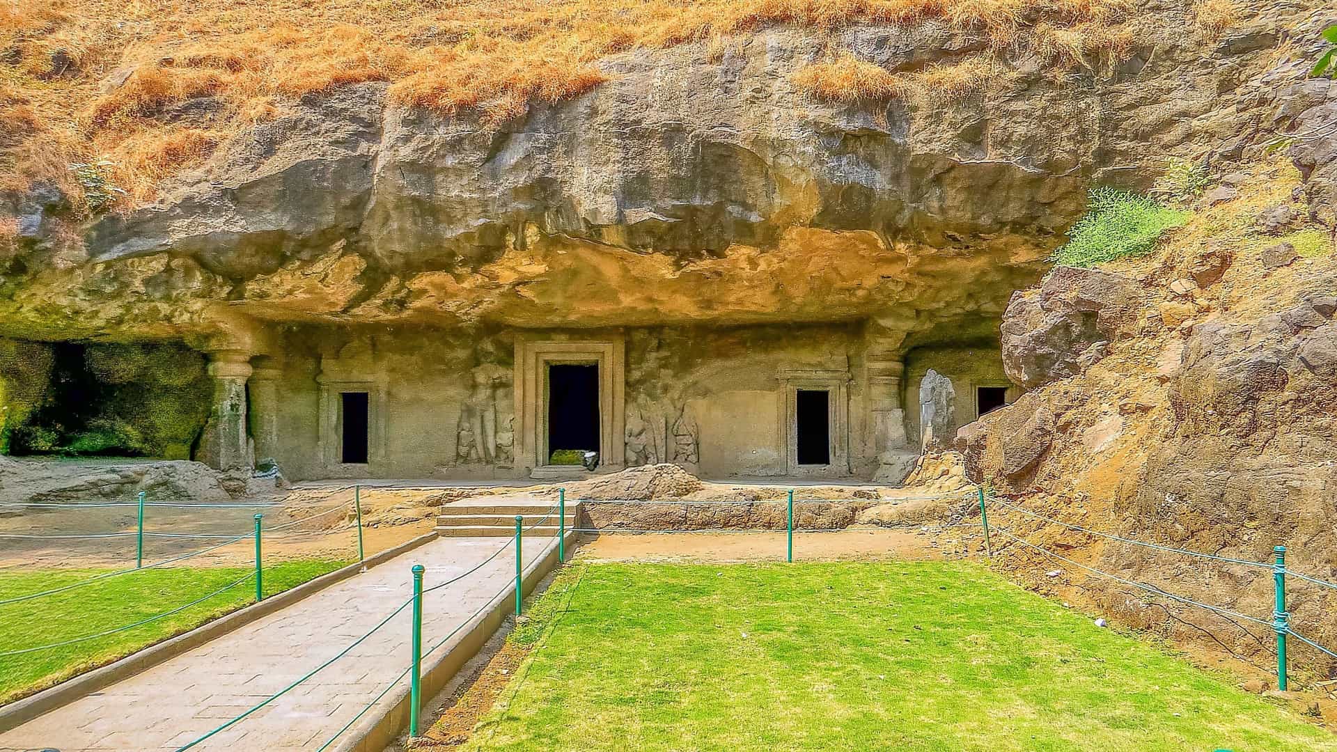 A wide-angle interior view of the Elephanta Caves, a UNESCO World Heritage site near Mumbai, showcasing the magnificent ancient rock-cut architecture dedicated to the Hindu god Shiva. The image features colossal stone pillars supporting the cavernous hall and highlights a beautifully preserved, intricately carved relief panel of a deity, demonstrating the extraordinary skill of 6th-century Indian artistry and religious devotion.