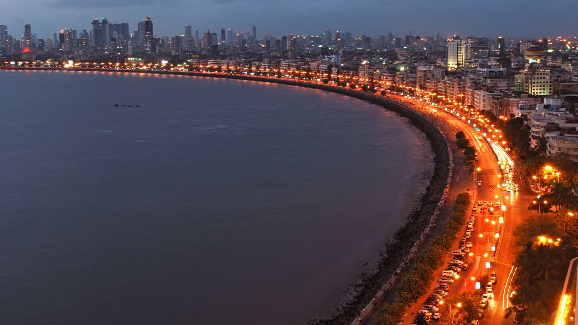 A high angle, panoramic night view of Marine Drive in Mumbai, India, famously known as the Queen's Necklace. The crescent shaped road curves along the Arabian Sea, with its streetlights illuminating the shoreline and resembling a sparkling string of pearls. The image shows the city's skyline with multiple high rise buildings and light trails from moving traffic.