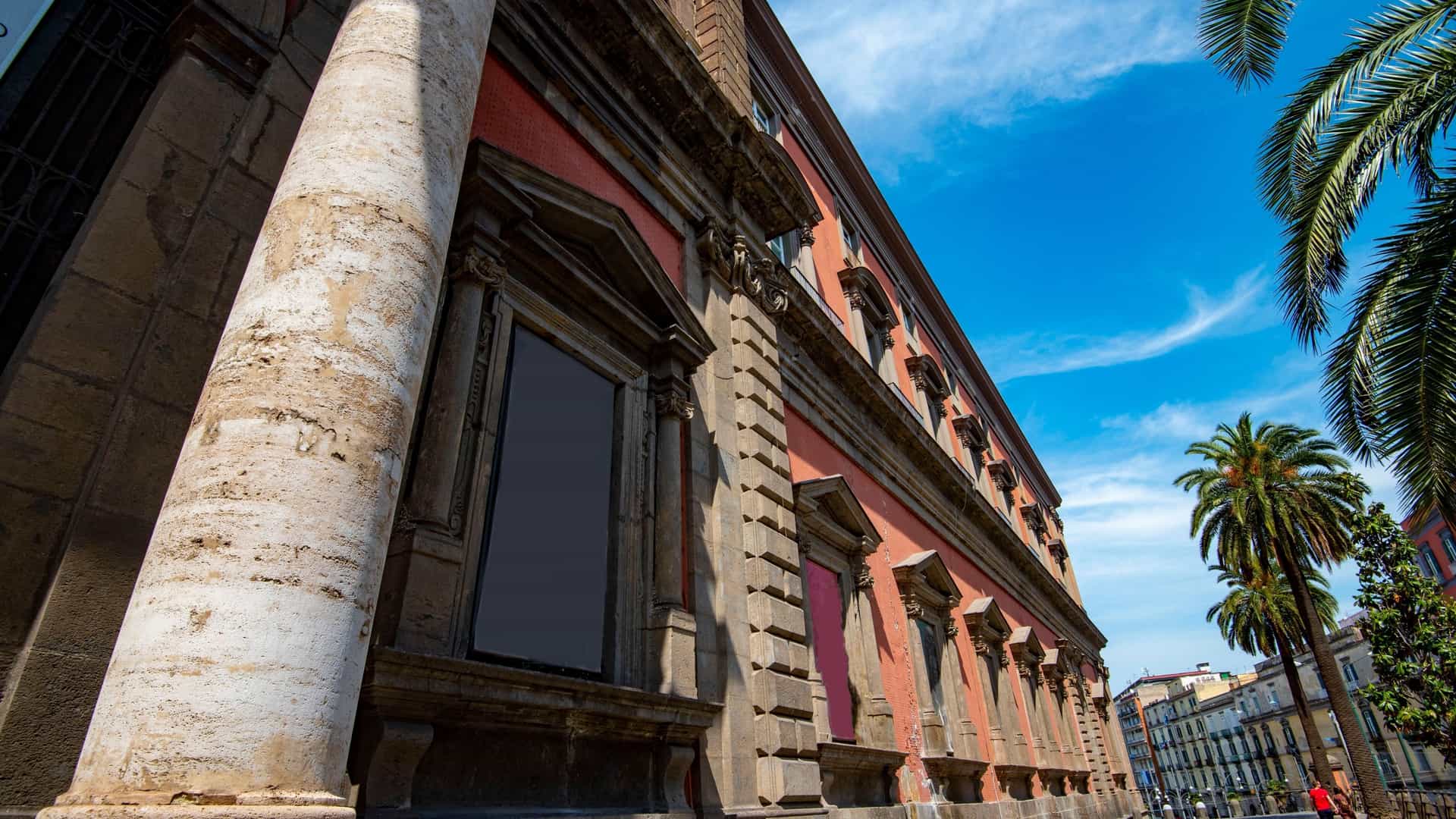 The exterior of the Naples National Archaeological Museum (MANN), a large, historic building in Naples, Italy. The neoclassical architecture features a series of arches and columns. The facade is a pale color, and the museum's grand entrance is visible. The photo captures the details of the building's architecture under a clear blue sky.