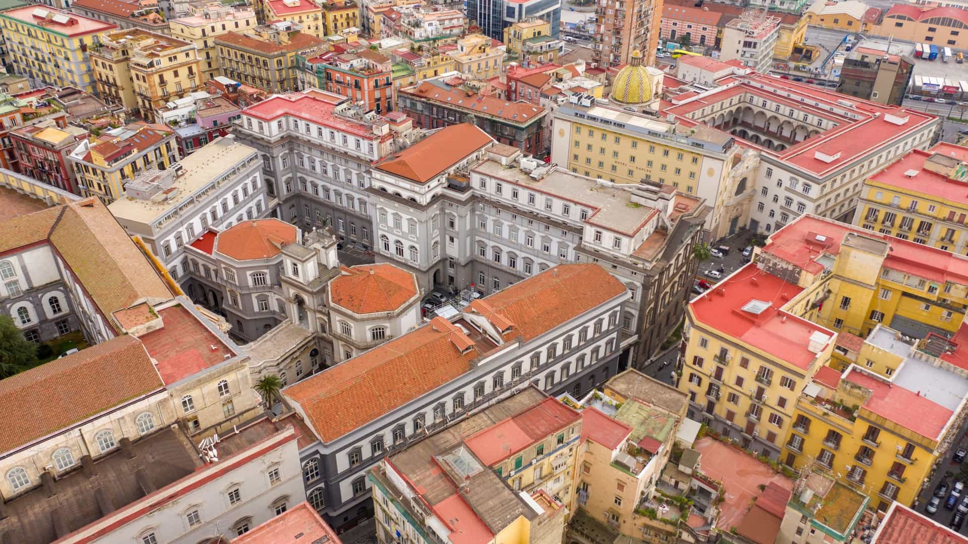 An aerial view of the historic city of Naples, Italy. The photograph shows a densely populated urban area with many buildings, including the historic center. In the distance, the Bay of Naples and Mount Vesuvius are visible under a clear sky, providing a panoramic view of the city and its famous natural surroundings.