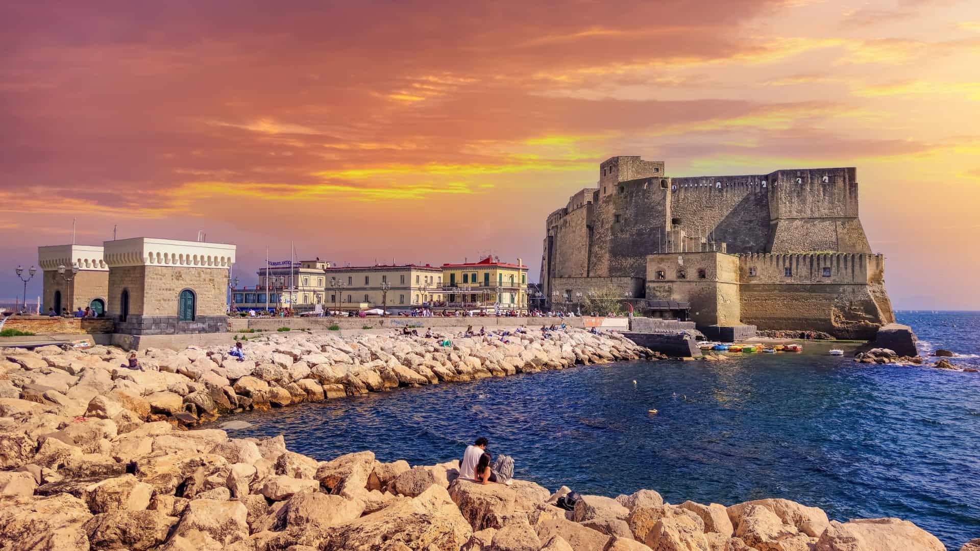 A scenic view of Castel dell'Ovo, a historic stone castle situated on a small peninsula in the Bay of Naples, Italy. The medieval fortress, with its imposing towers and walls, is connected to the mainland by a long causeway. The image captures the castle on the calm blue water, with the city of Naples and its coastline visible in the background under a bright sky.
