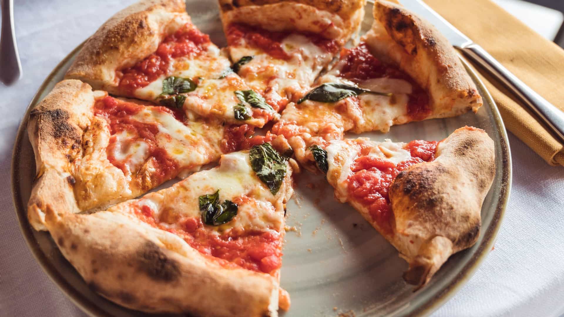 A close-up photograph of a classic Margherita style pizza, demonstrating the simple perfection of a Pizza at L'Antica Pizzeria da Michele. The large, round pie features a thin, slightly wet center topped with bright red tomato sauce, fresh mozzarella cheese, and scattered basil leaves, all framed by a high, blistered, and lightly charred cornicione (crust).
