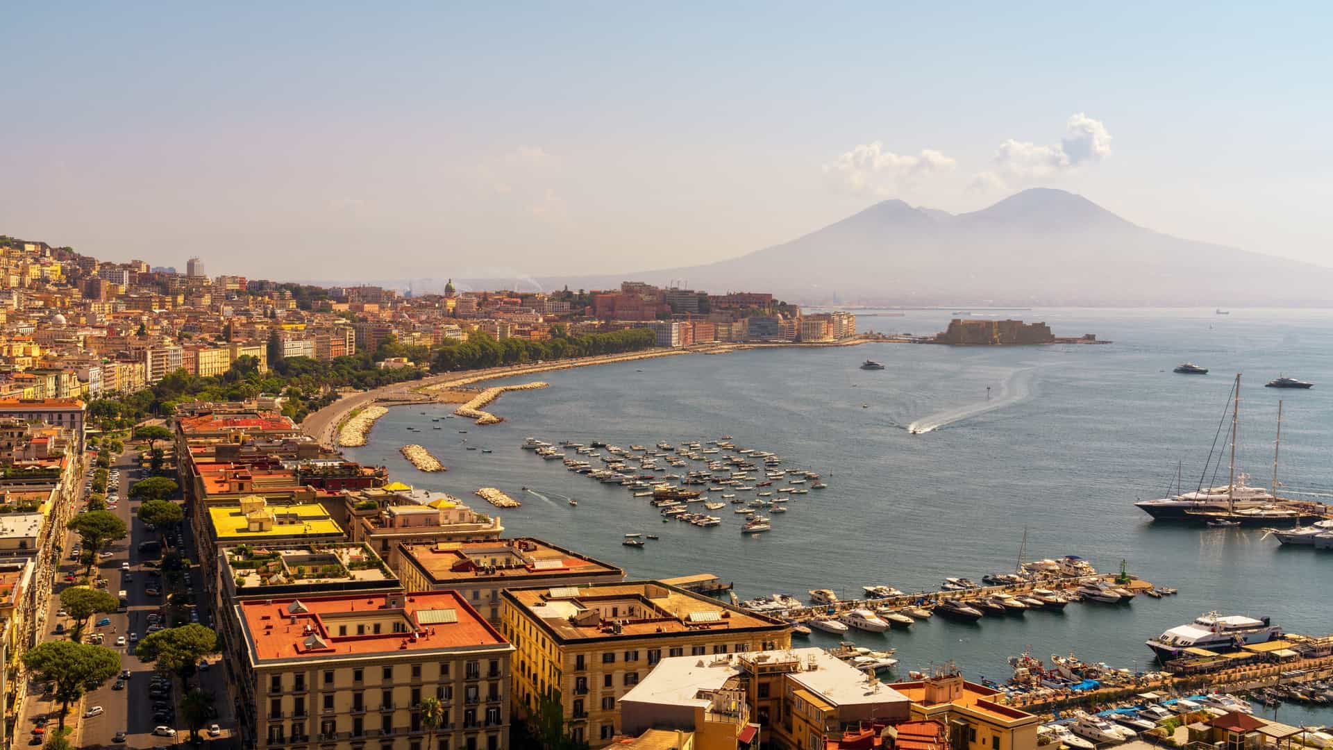 A wide panoramic view of Naples, Italy, showcasing the densely packed cityscape along the harbor, the calm blue water of the Gulf of Naples, and the iconic, massive cone of Mount Vesuvius dominating the background horizon.
