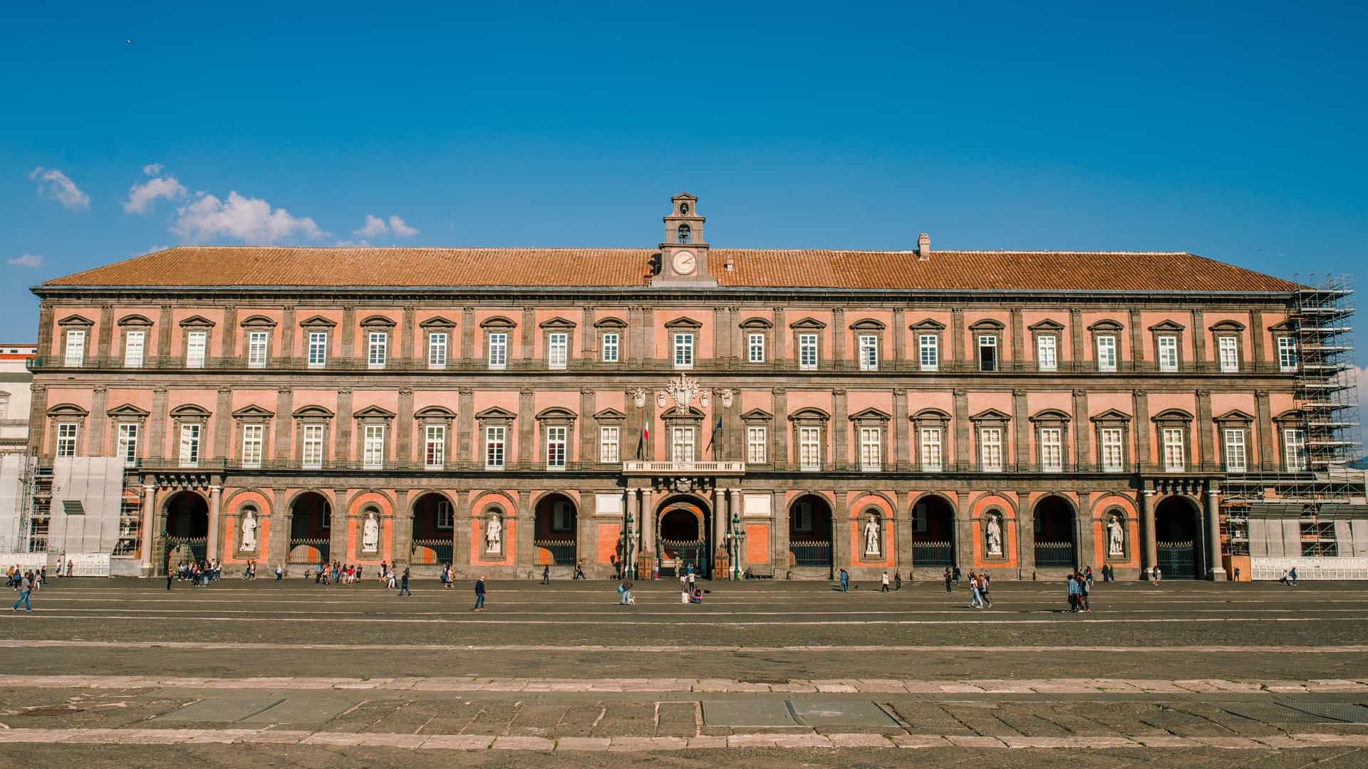 The Palazzo Reale di Napoli (Royal Palace of Naples) in Naples, Italy. The photograph captures the imposing Baroque and Neoclassical facade of the palace, which faces the city's main public square, Piazza del Plebiscito. The building's exterior is characterized by its red and grey stone and a series of large arched windows and a portico, with the grand architecture reflecting its history as a royal residence.