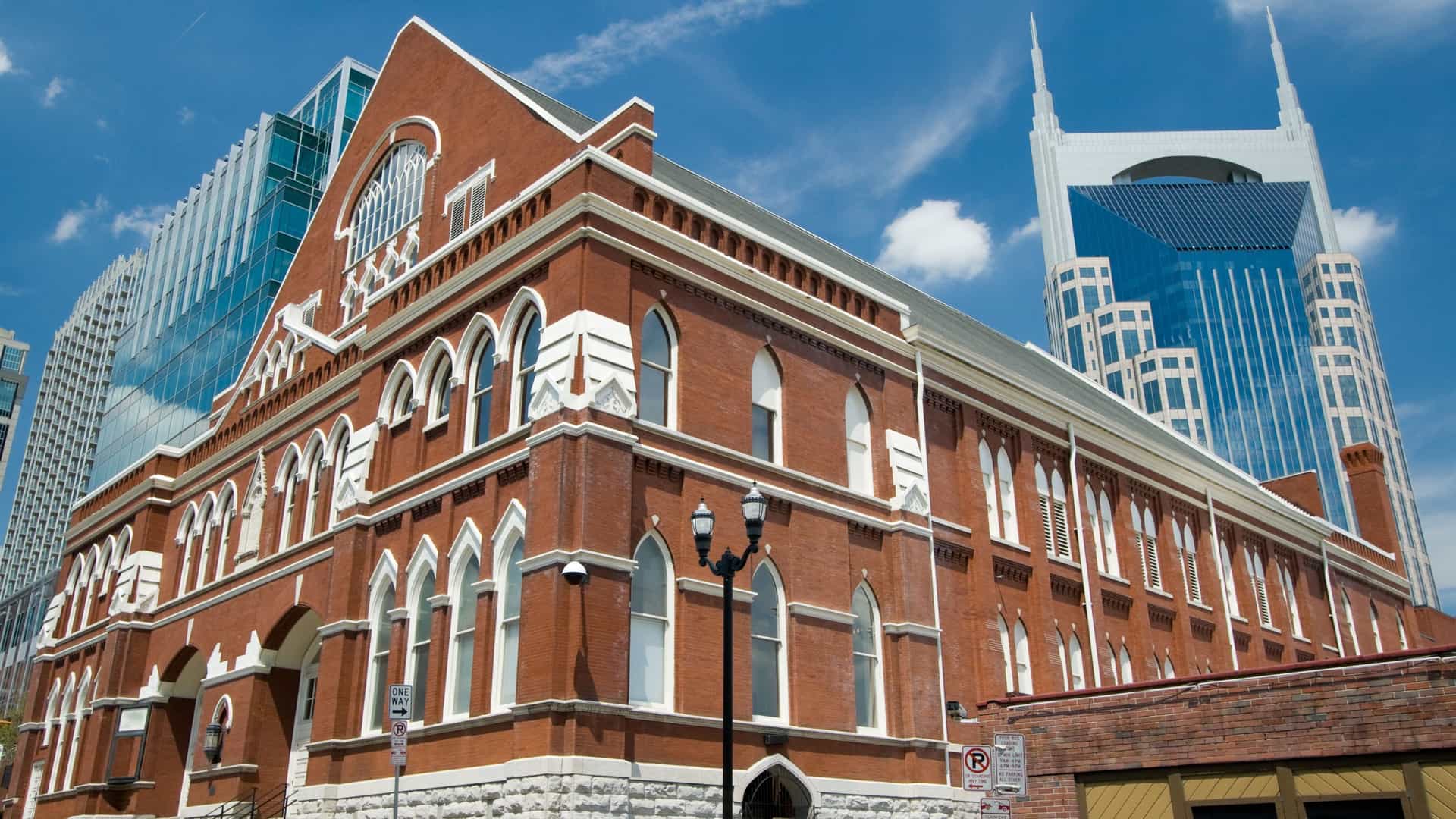 The Ryman Auditorium, a historic red brick building in Nashville, Tennessee. The entrance features white signage with bold lettering, reading 'RYMAN AUDITORIUM' and often mentioning its nickname 'Mother Church of Country Music'