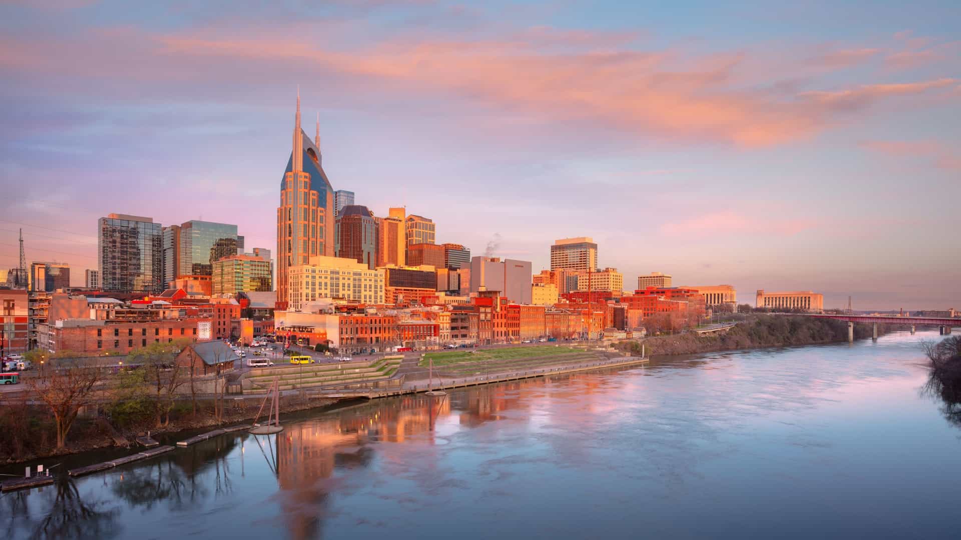 A panoramic view of the downtown Nashville, Tennessee cityscape. The image features the skyline with its prominent skyscrapers, including the notable AT&T building, reflected in the Cumberland River. The city's bridges and a clear sky complete the scene, which captures the urban landscape of Music City.