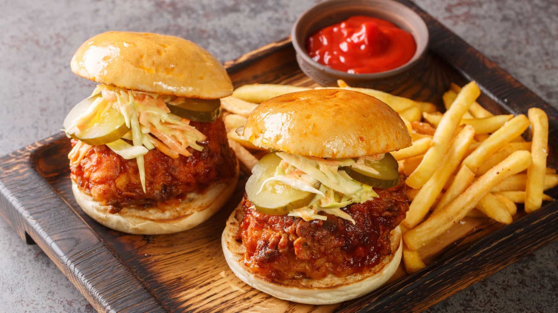 A piece of Nashville Hot Chicken, a deep-fried chicken thigh or breast, slathered in a vibrant red, spicy cayenne-based oil and served atop a slice of white bread. The dish is garnished with several bright green pickle slices.