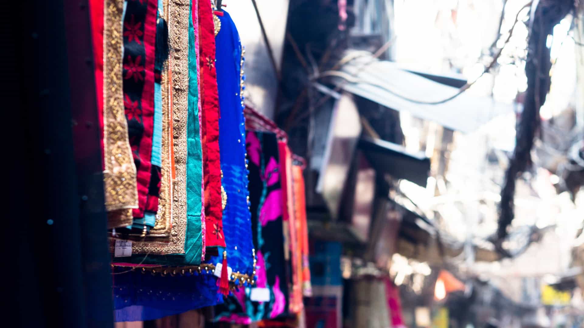 A close-up view of numerous beautiful, colorful handicraft scarves and shawls hanging on display at a street-side market stall, likely in a city like Delhi, India. The textiles feature a mix of colors and patterns, suggesting they are a popular item for sale.