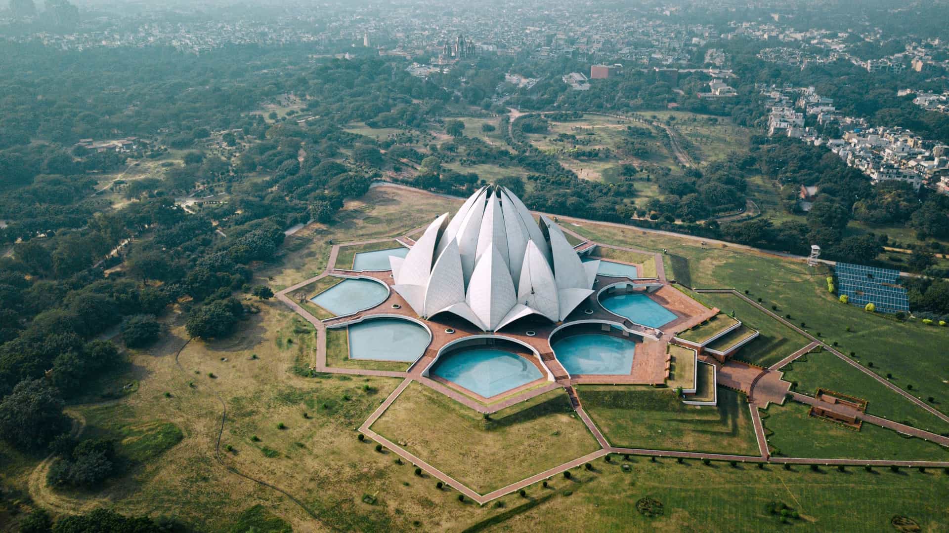 An aerial view of the Lotus Temple in New Delhi, India. The modern Baháʼí House of Worship is a white, marble clad structure designed in the shape of a lotus flower, with clusters of free standing "petals" forming nine sides. The temple is surrounded by lush green lawns and a symmetrical arrangement of pathways and pools.