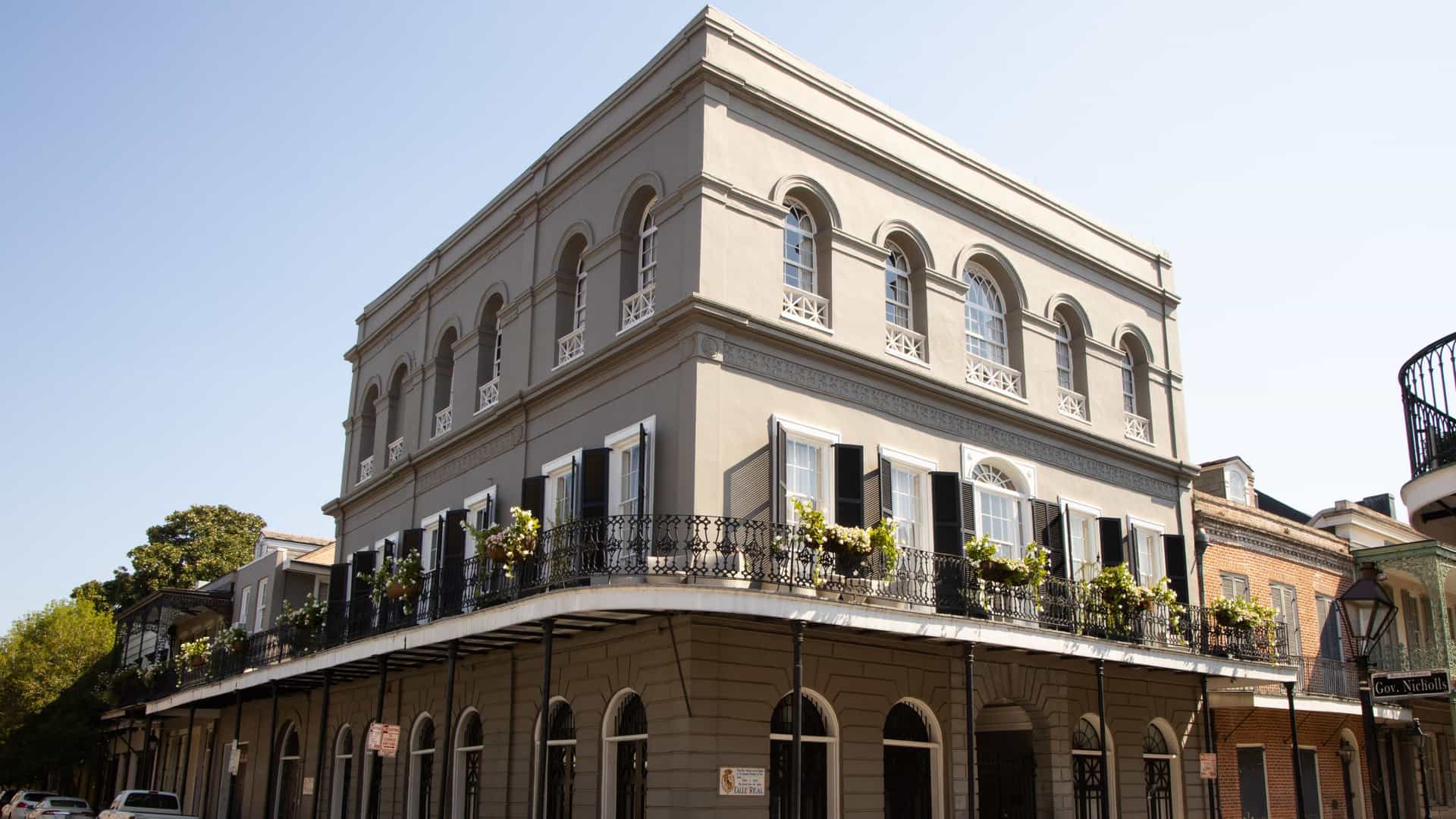 A large, historic corner building on Royal Street in New Orleans, Louisiana, featuring a gray stucco facade and three upper stories with arched windows. The ground floor has a continuous arcade, and a multi-level black cast-iron balcony wraps around the corner, covering the sidewalk below. The street corner is visible in the foreground under a clear, sunny sky.