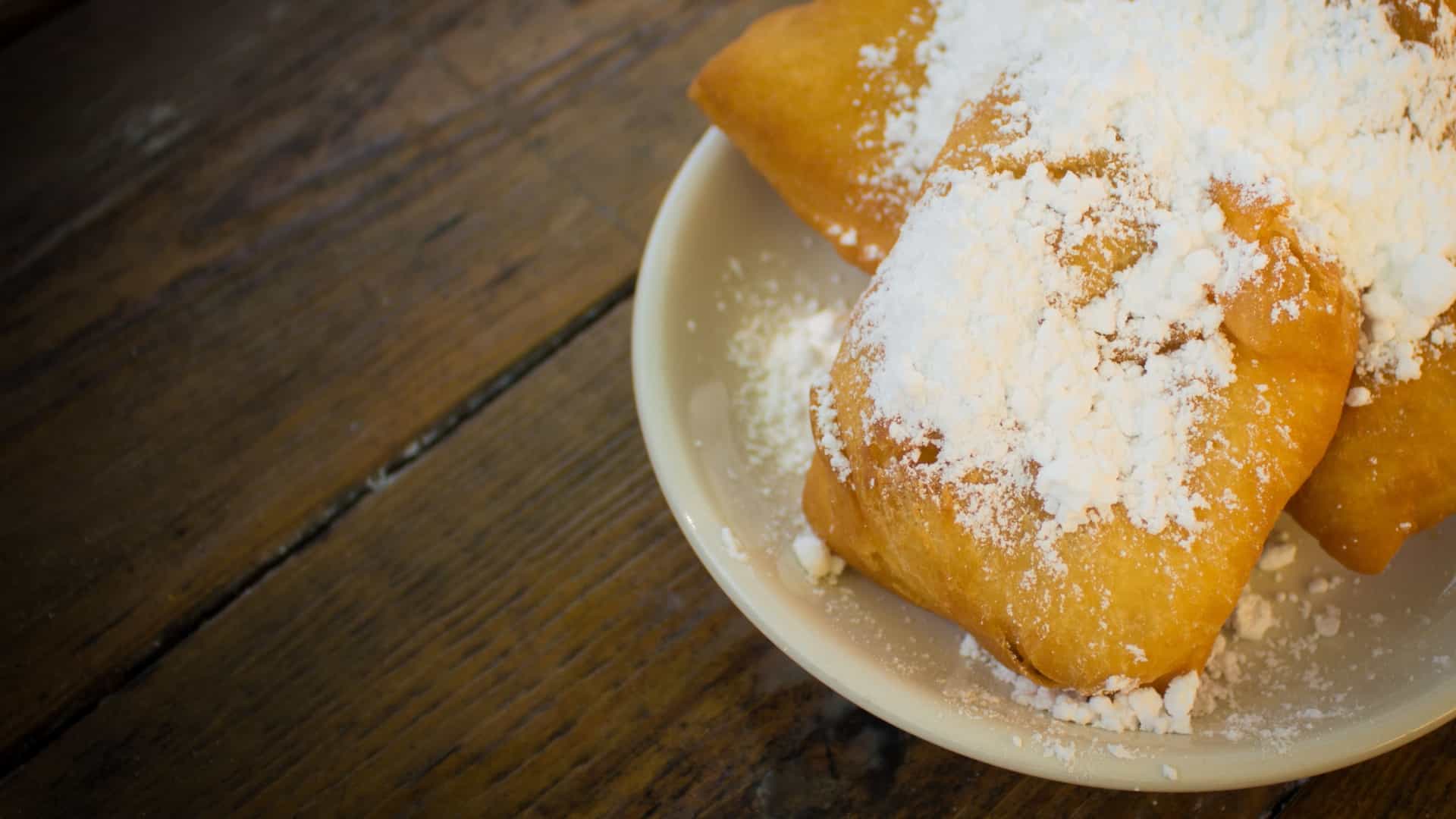 A close up of a plate of beignets, a classic New Orleans dessert associated with Café du Monde. The square pieces of deep fried dough are generously covered in powdered sugar and are resting on a white plate, which is set on a rustic wooden table.