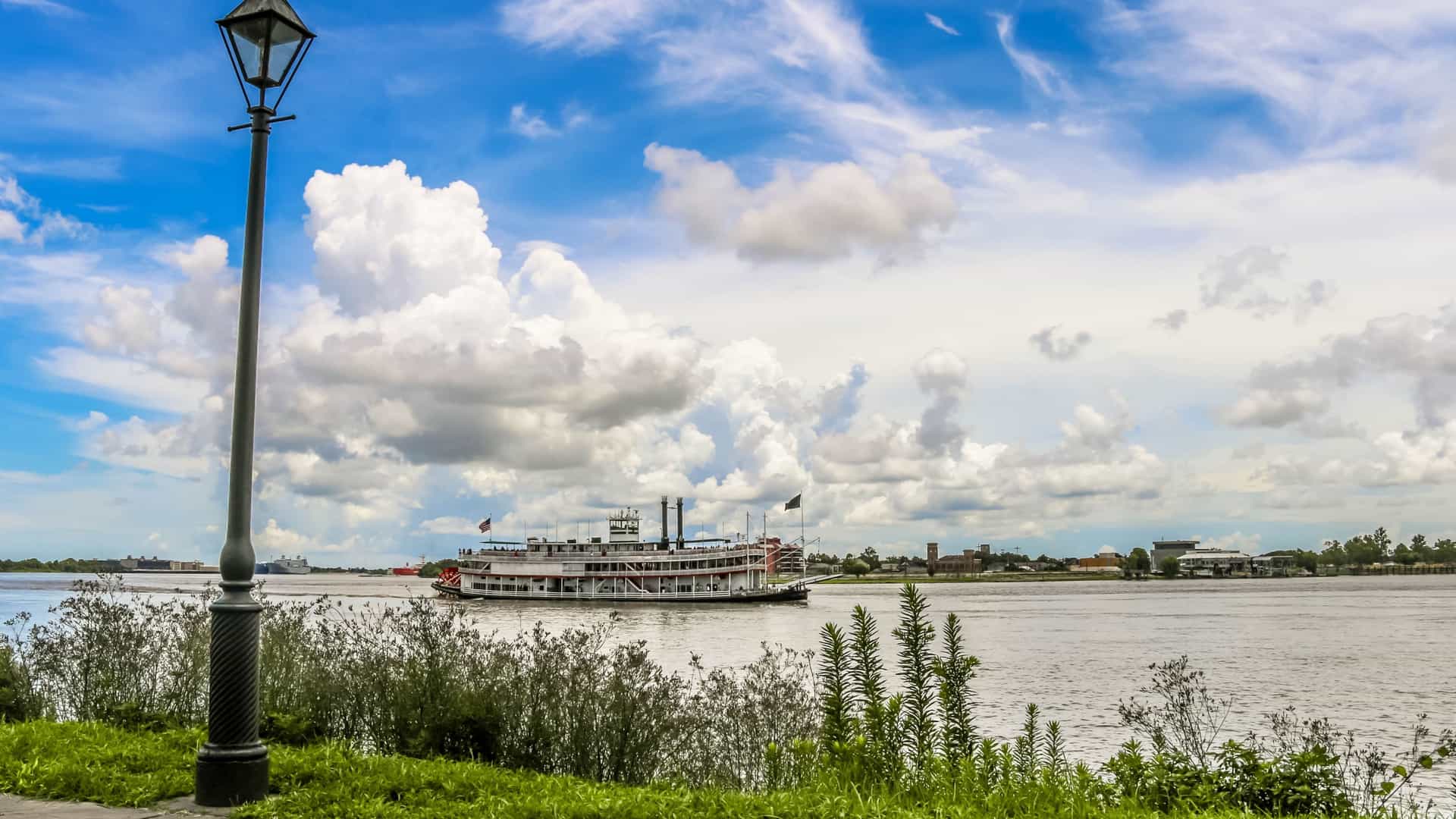 A steam powered paddle wheeler boat traveling on a wide, calm river, most likely the Mississippi. The boat has multiple decks and a large red paddle wheel at the stern, creating a sense of a bygone era of river travel.