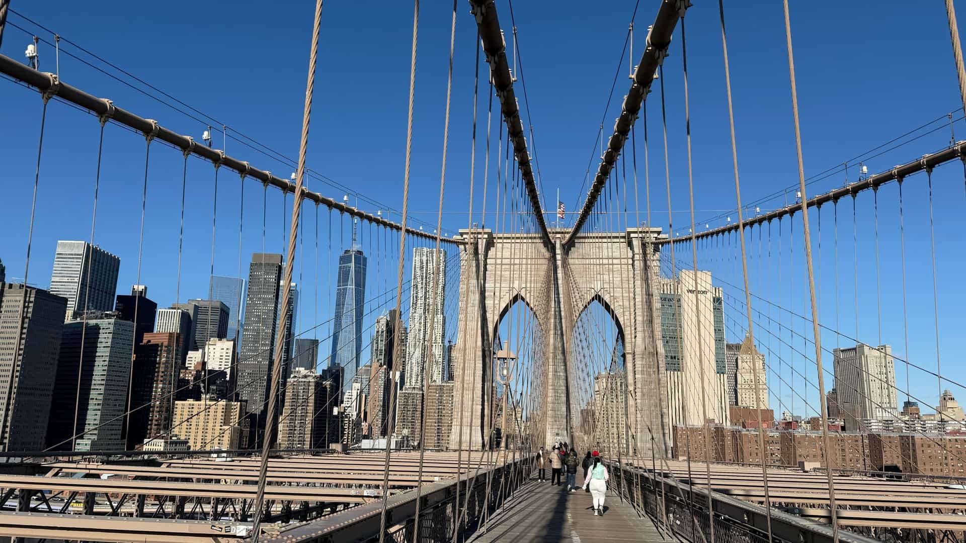 A person is walking across the Brooklyn Bridge on a sunny morning. The image shows the bridge's wooden pedestrian walkway, the distinctive Gothic-arch towers, and the steel cables that support the structure. The Manhattan skyline is visible in the background.