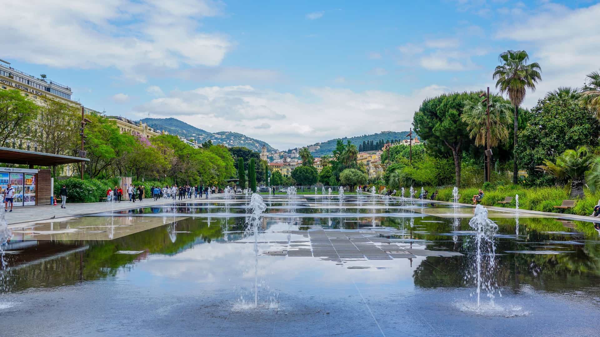 A panoramic view of the Promenade du Paillon, a large green urban park in Nice, France. The image shows the park's landscaped areas, including a water mirror with jets, surrounded by walkways and lush trees. The city's buildings and the famous Place Masséna are visible in the background.