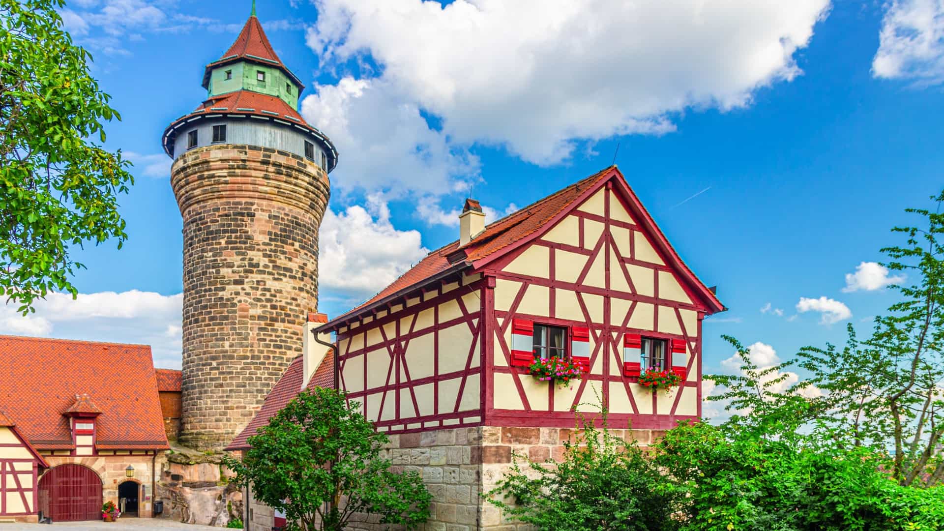 A close up, low angle view of the medieval stone walls and cylindrical Sinwell Tower of the Nuremberg Castle in Germany, with the castle's red roofed buildings and towers visible on a sandstone ridge in the background. The castle is framed by a clear blue sky.