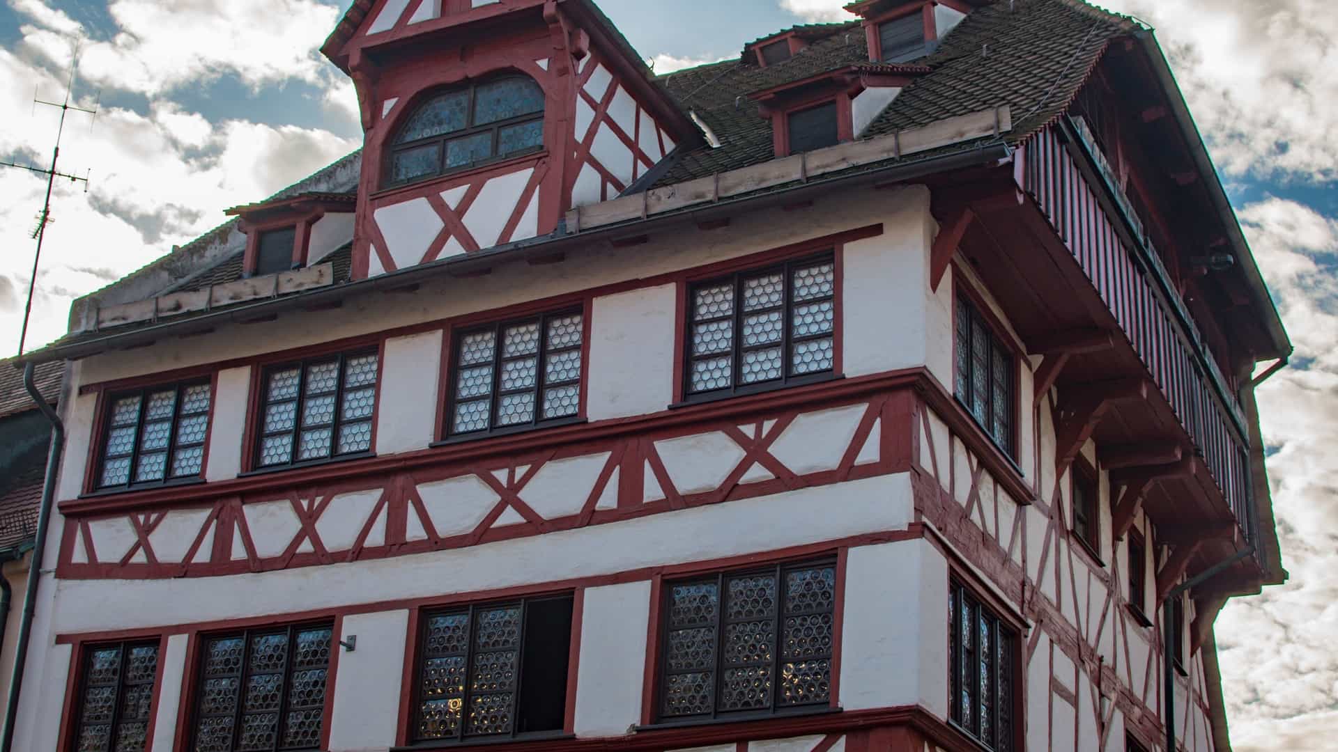 A view of Albrecht Dürer's House in Nuremberg, Germany, a five story building with a red tiled half hip roof. The top three floors are made of dark half timbered wood with white infill, while the bottom two floors are made of light colored stone. The building is situated on a cobblestone street with a tree in front of it and other historic buildings nearby.