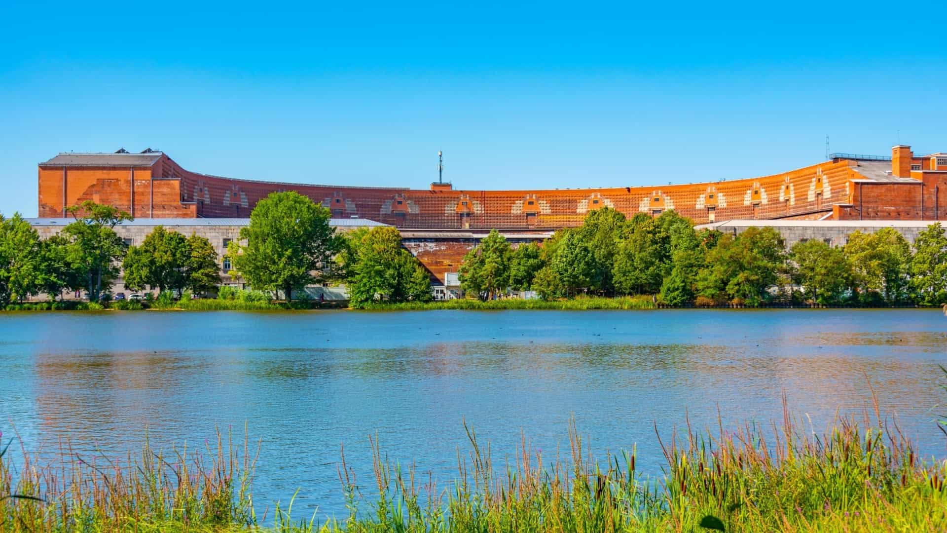 A wide-angle view across the Grosser Dutzendteich lake towards the colossal, brick Congress Hall (Kongresshalle) at the former Nazi Party Rally Grounds in Nuremberg, Germany, which also houses the Documentation Centre Nazi Party Rally Grounds. The distinctive, C-shaped red brick structure is visible above a row of green trees, contrasting sharply with the bright blue sky, with tall grasses and reeds visible in the foreground near the water's edge.
