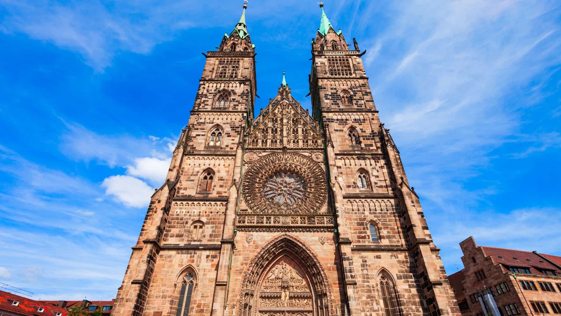 An exterior view of the towering St. Lorenz (Saint Lawrence) Church in Nuremberg, Germany, a magnificent example of medieval Gothic architecture. The image shows the church's intricate facade with its large rose window and twin spires, rising above the surrounding buildings in the city center.